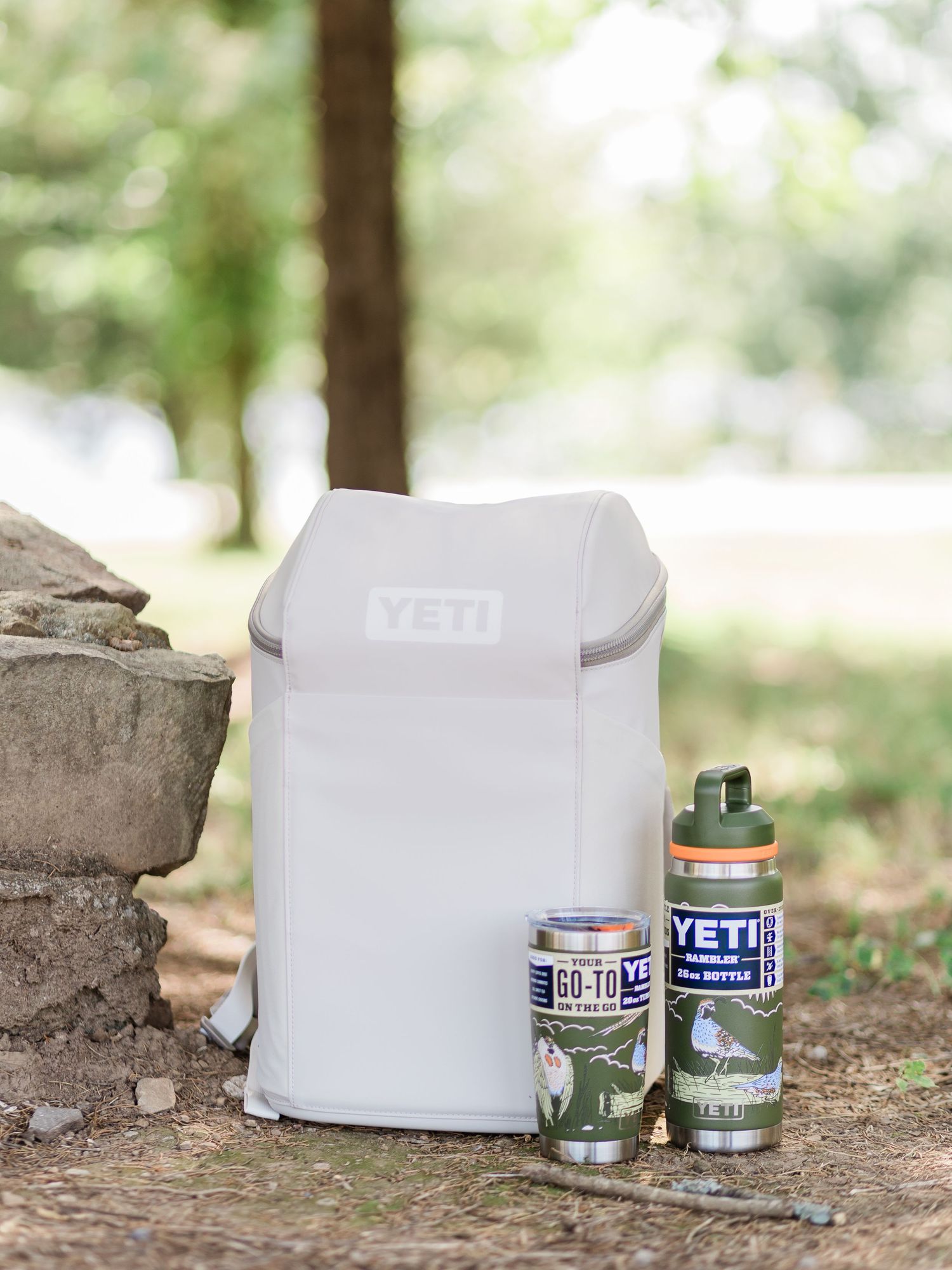 YETI backpack cooler with Rambler tumbler and water bottle displayed outdoors on stone surface in natural setting.