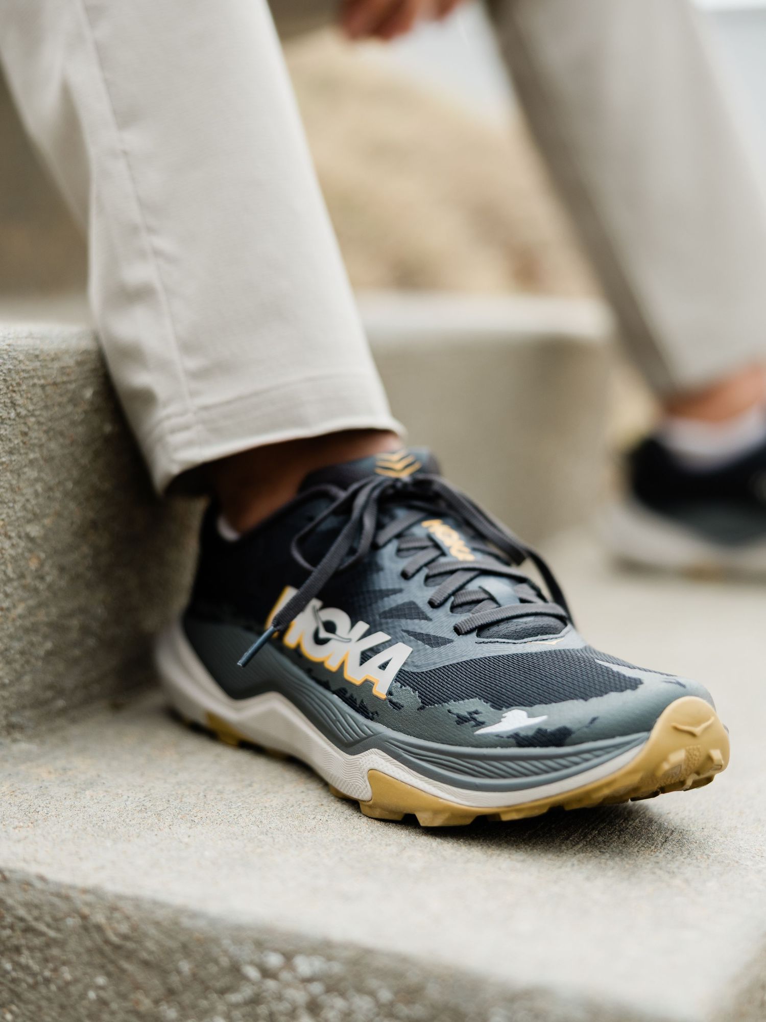 HOKA trail running shoes in dark gray and gold colorway worn on concrete steps with white pants.