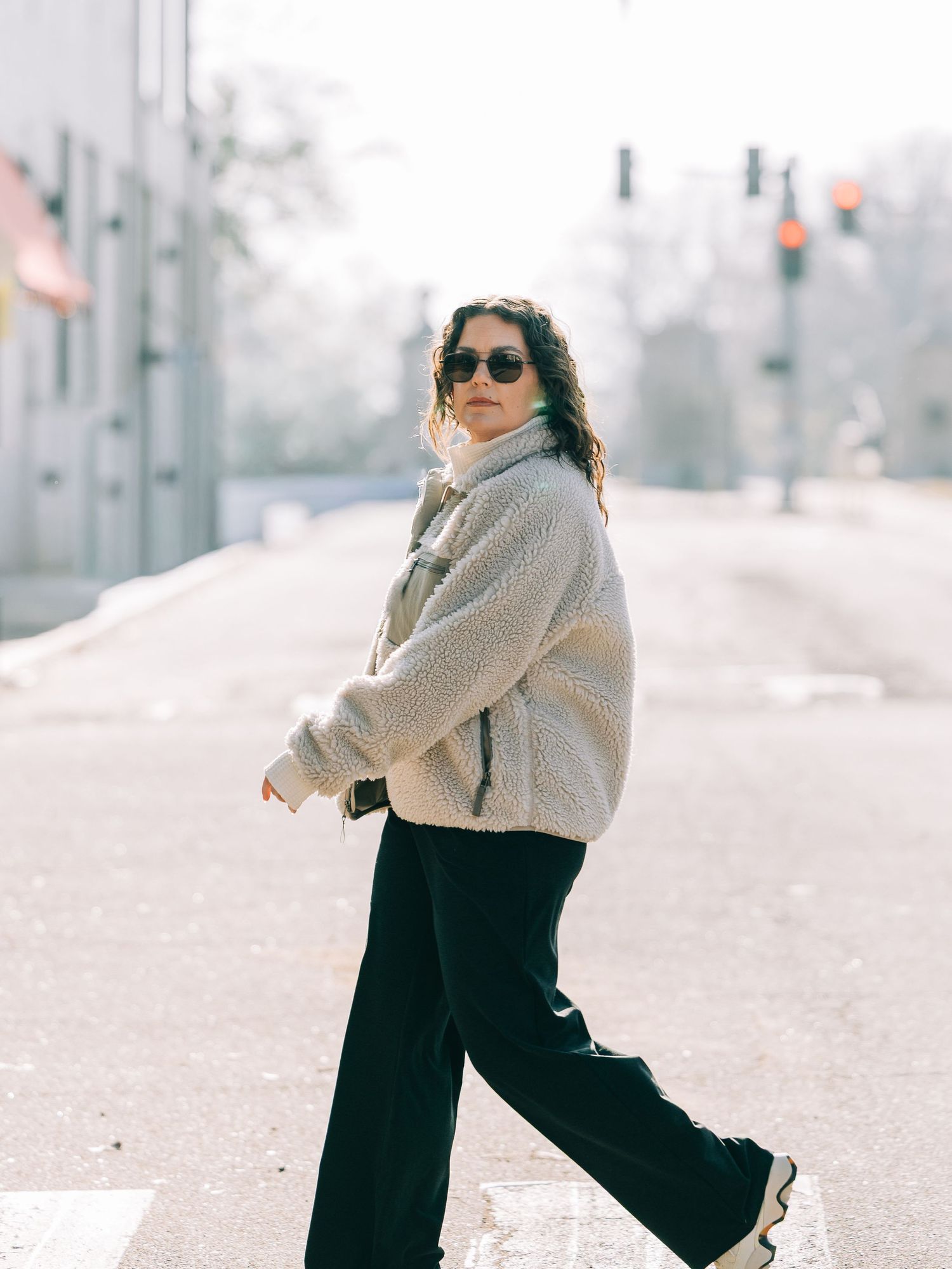 Stylish winter outfit with beige teddy coat, black wide-leg pants and sunglasses on a misty urban street.