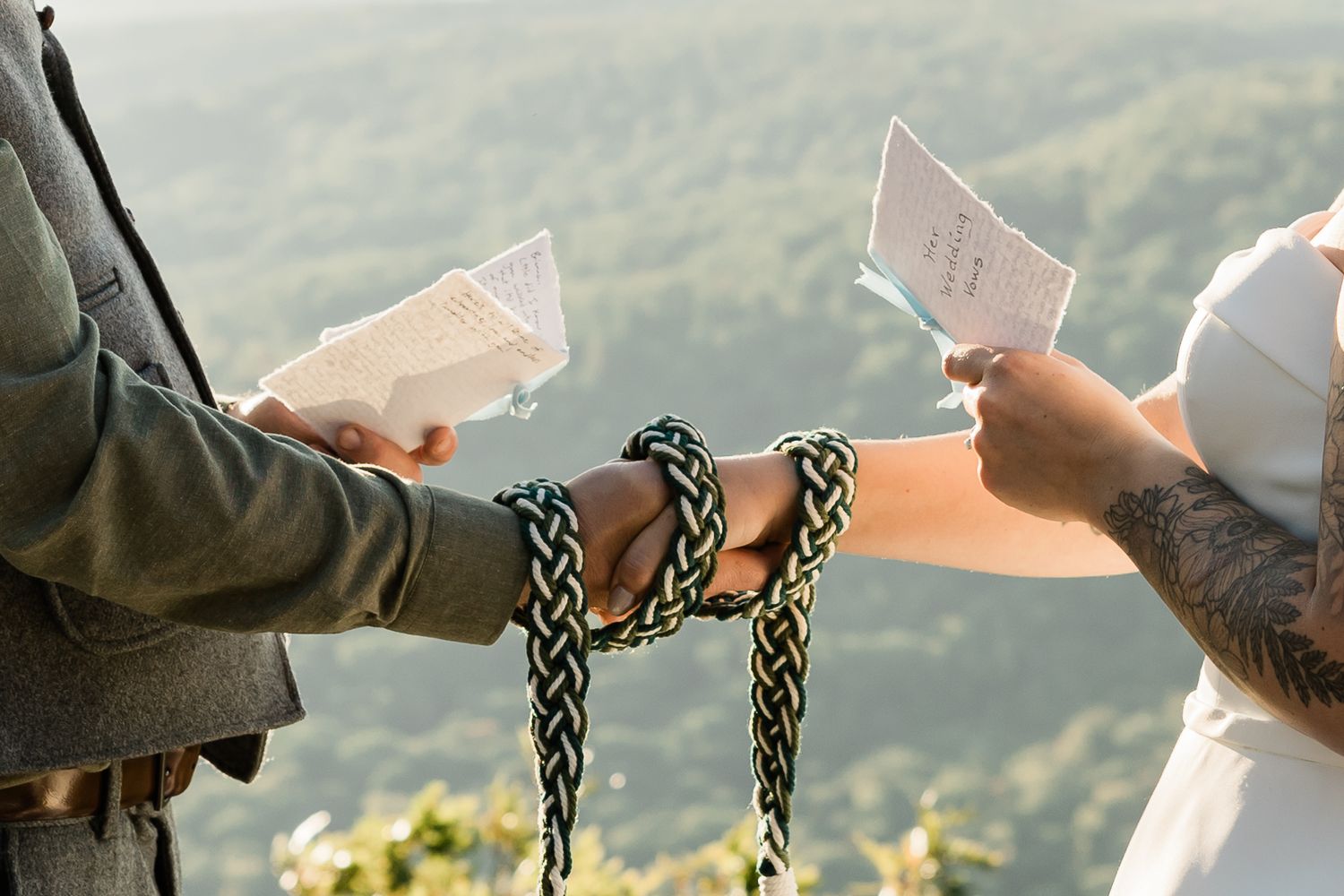 Close-up view of hands performing a traditional handfasting ceremony with braided cords during an outdoor wedding ritual.