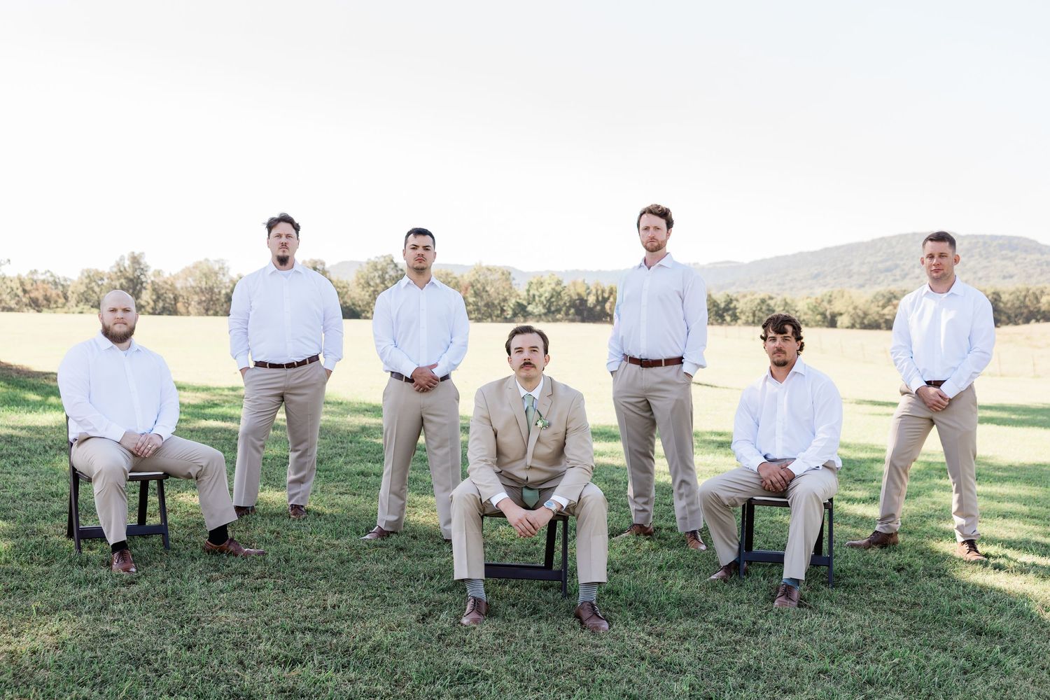 Wedding party members in khaki pants and white shirts sit casually on grass at an outdoor venue.