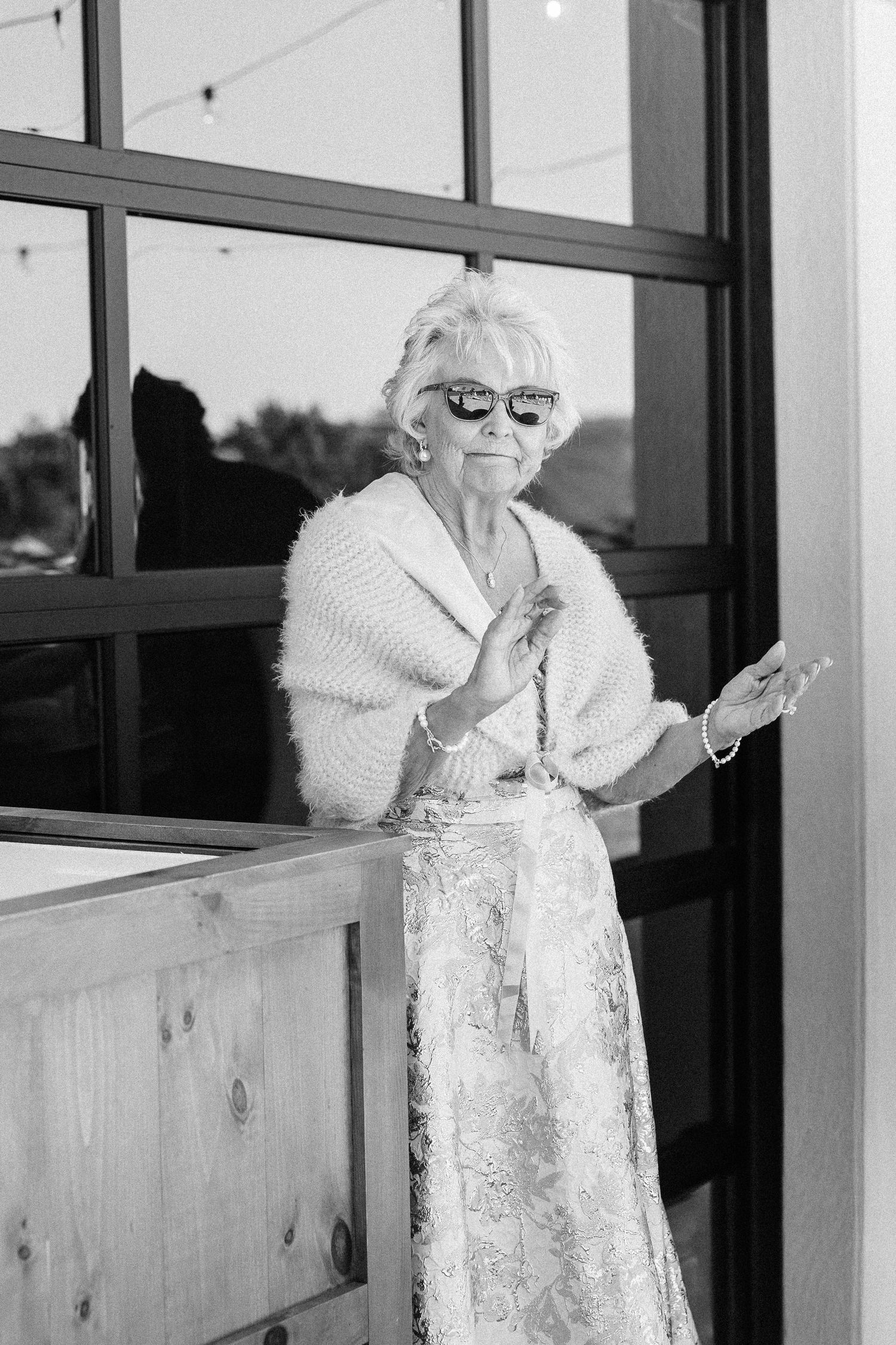 Elegant senior woman in white lace dress standing by glass windows.