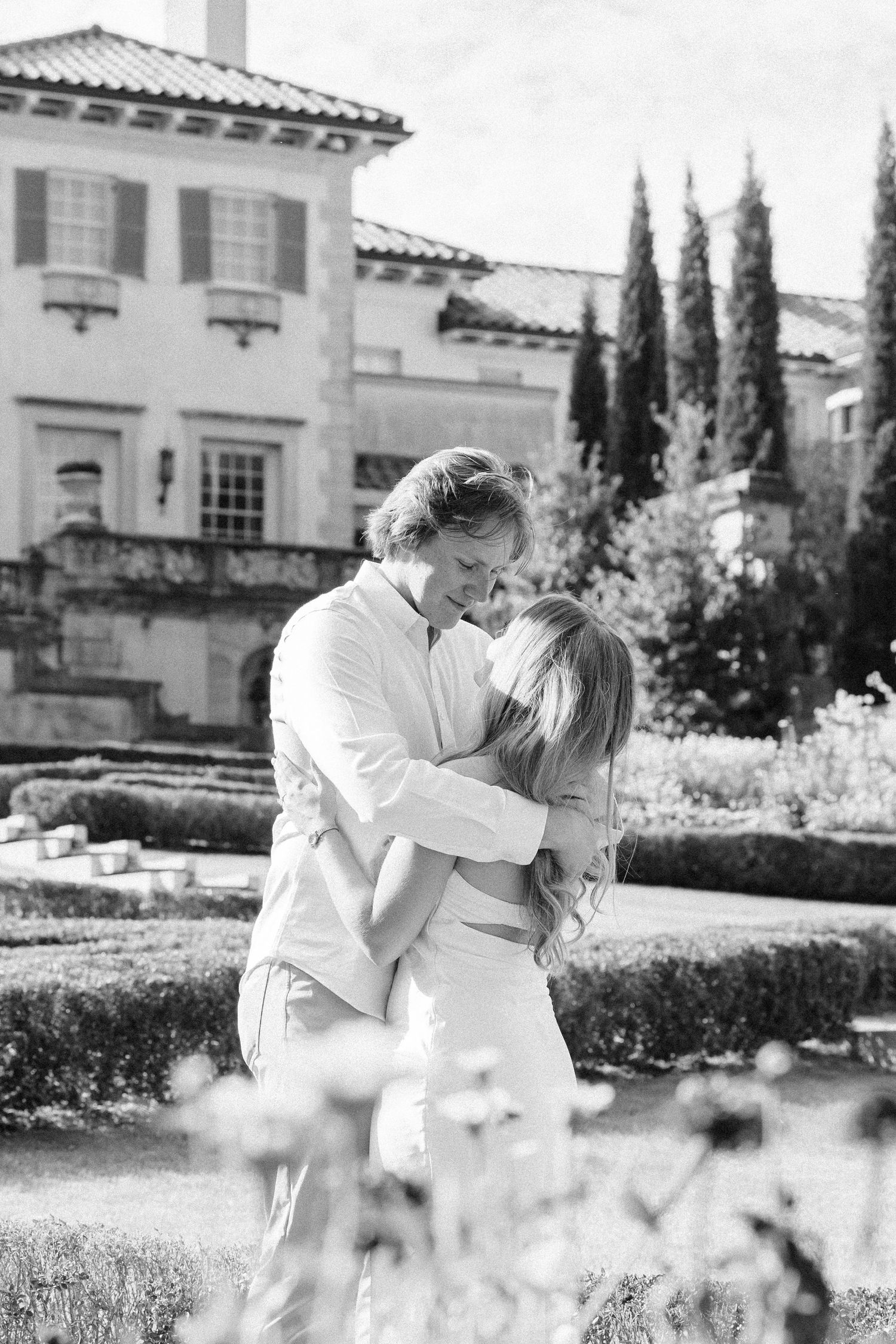 A romantic couple in white outfits embrace in a garden setting with Mediterranean-style architecture in the background.