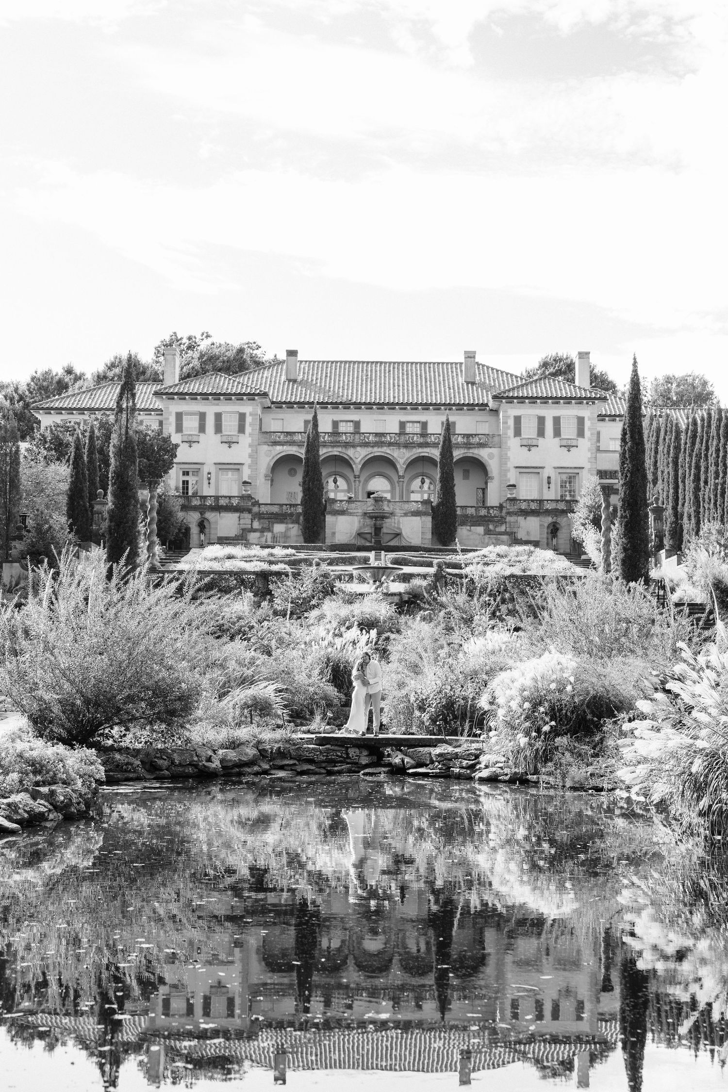 A grand Mediterranean-style mansion with arched windows reflects perfectly in a serene garden pond, captured in black and white.