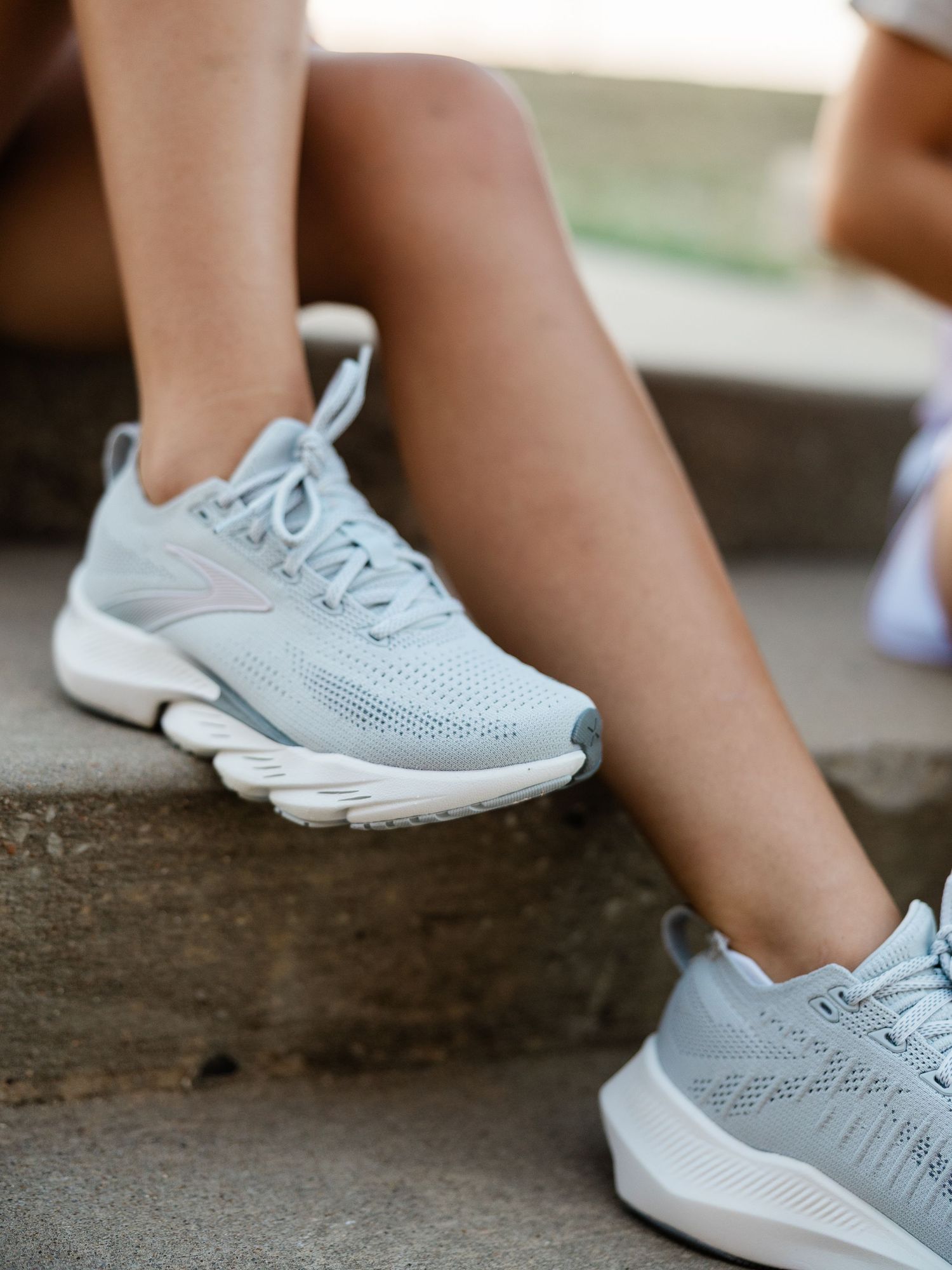 Woman sitting on stone steps wearing light blue Brooks running sneakers with white soles.