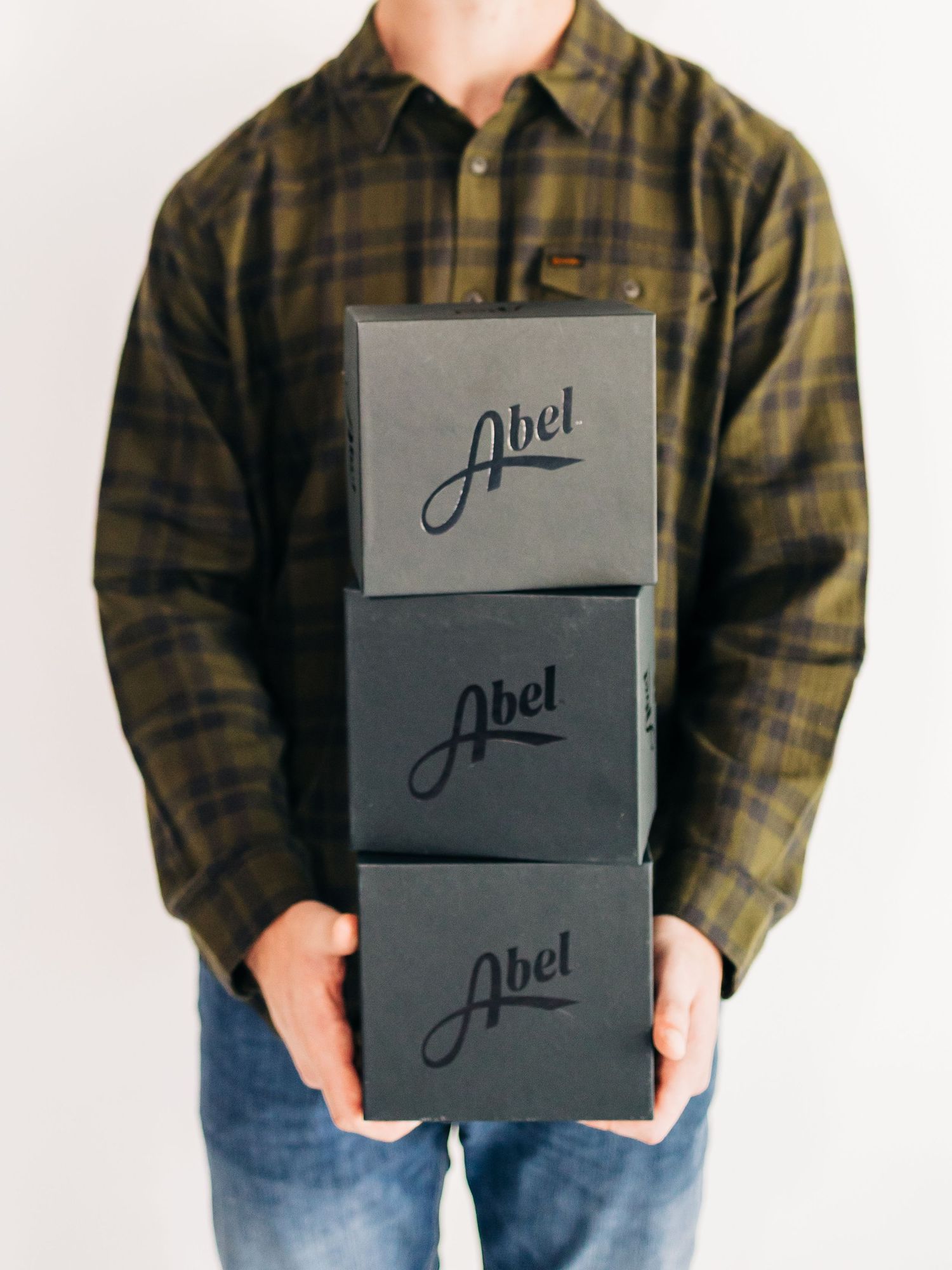 Person in plaid shirt holding stacked black gift boxes with 'Joel' branding against white background.