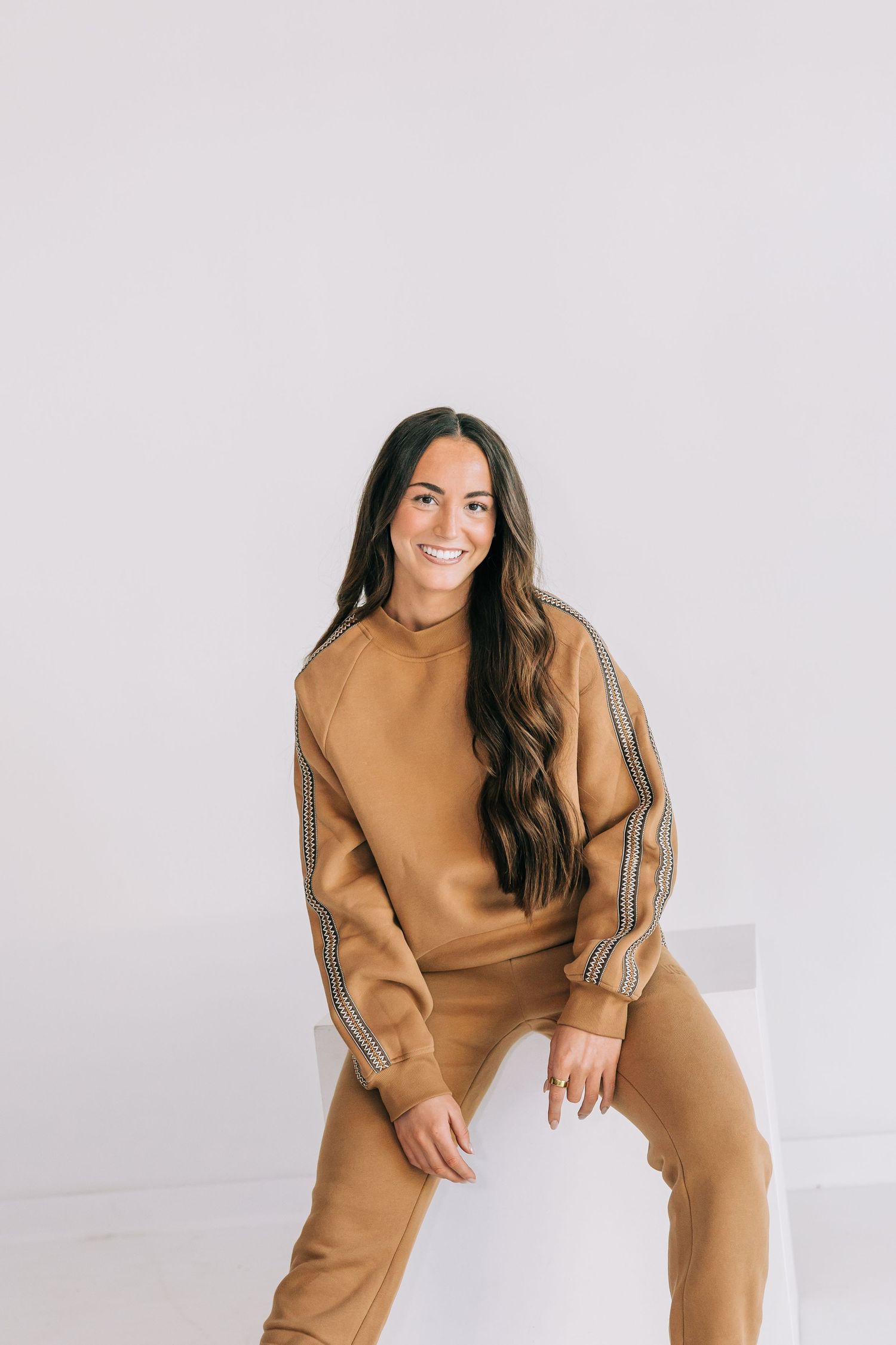 Woman in tan tracksuit lounges and poses casually on white background.