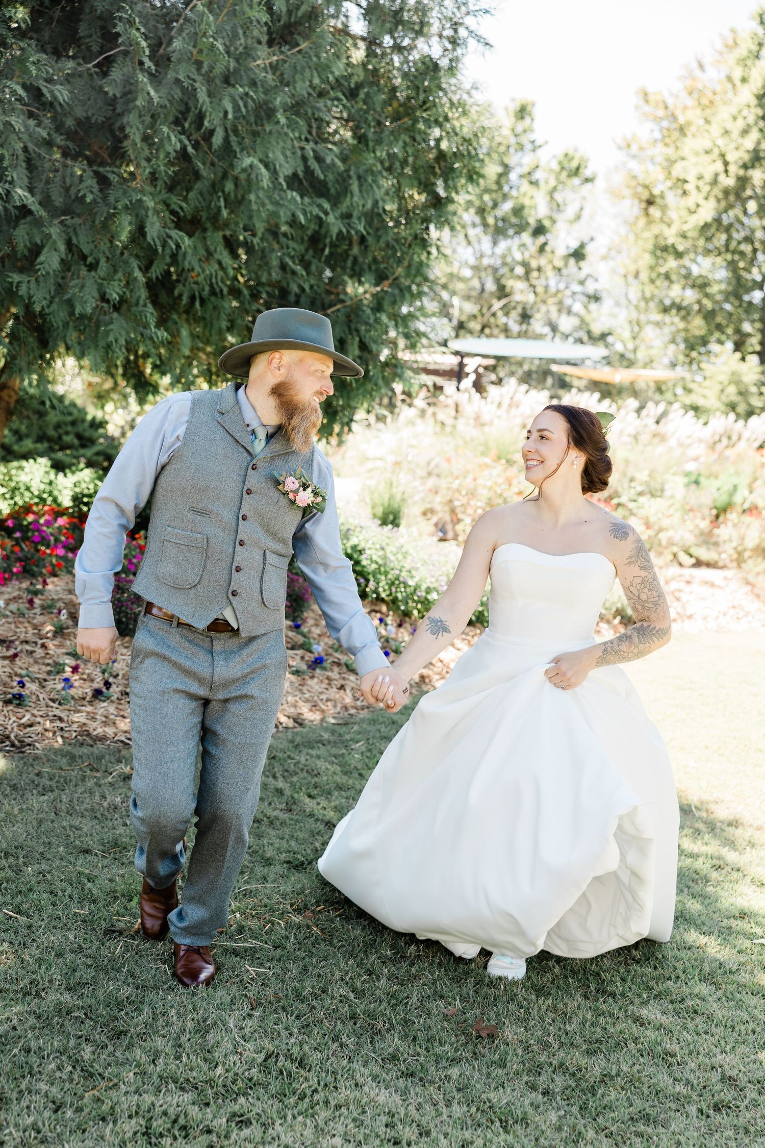 Joyful couple in wedding attire dance and twirl together on a grassy lawn surrounded by evergreen trees.