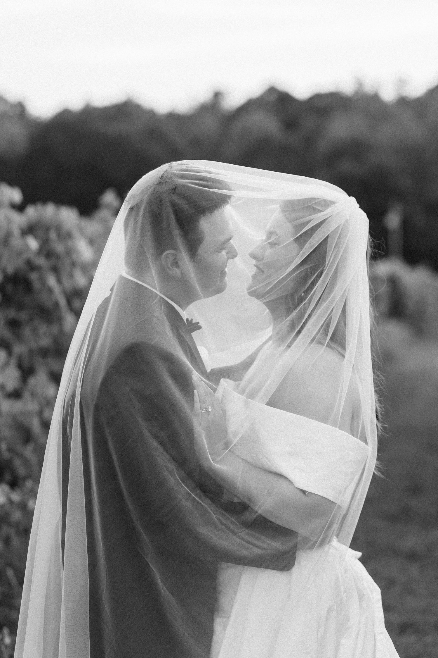 A romantic wedding couple embraces under a flowing white veil in a black and white outdoor portrait series.