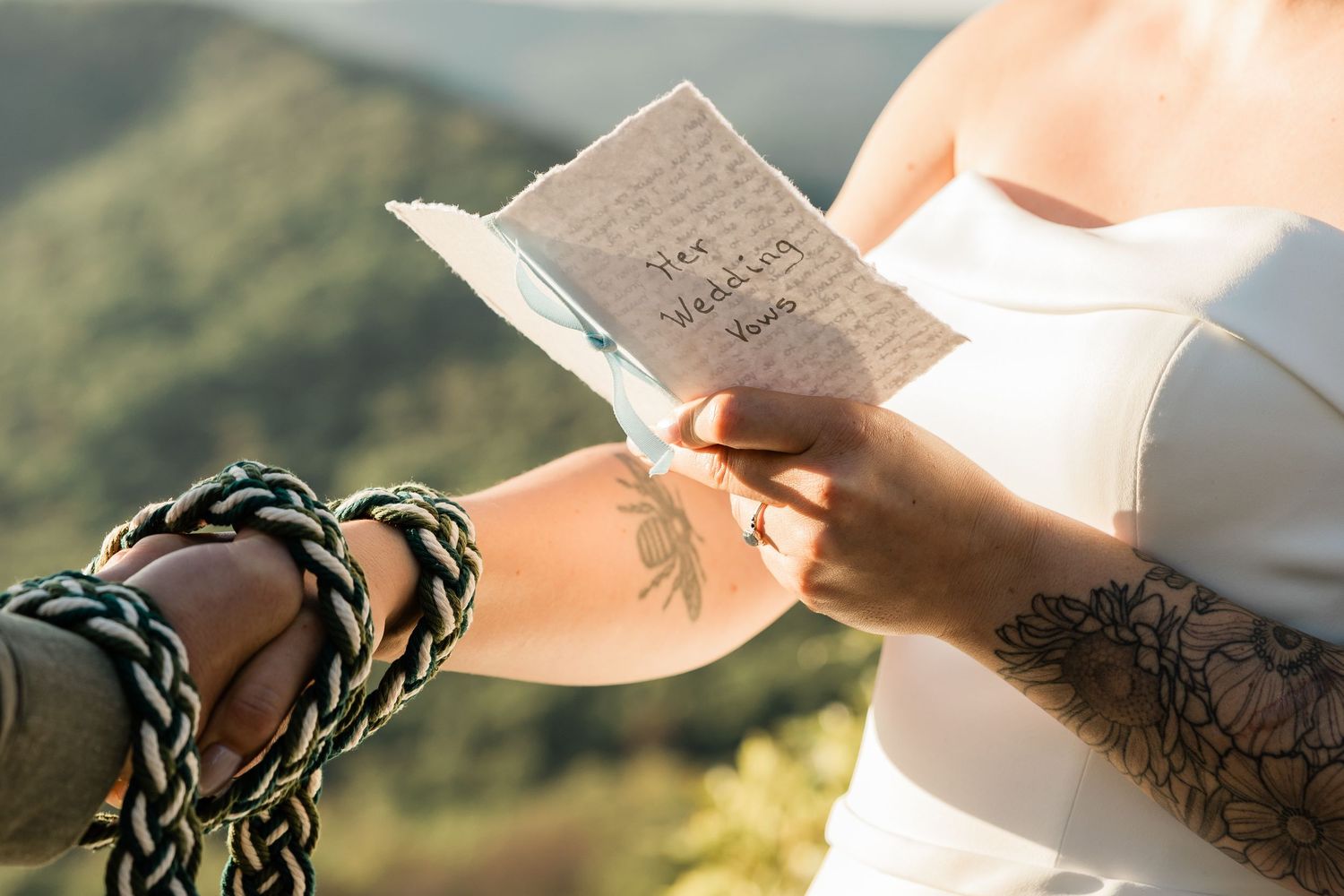 A couple wearing wedding attire stands close together against a scenic mountain backdrop during golden hour.