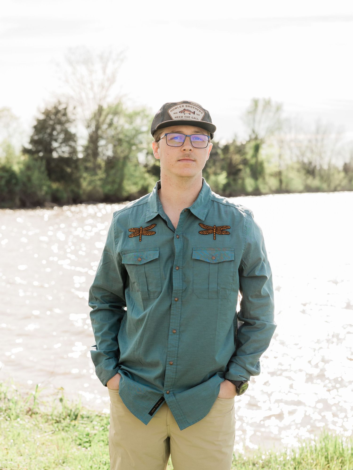 Young man in teal shirt and cap stands by a sunlit river with green trees in the background.
