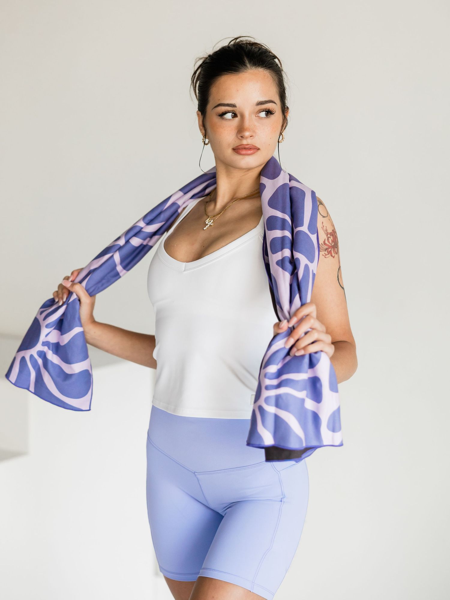 Woman in white tank top and lavender bike shorts wearing a purple patterned towel around her neck.