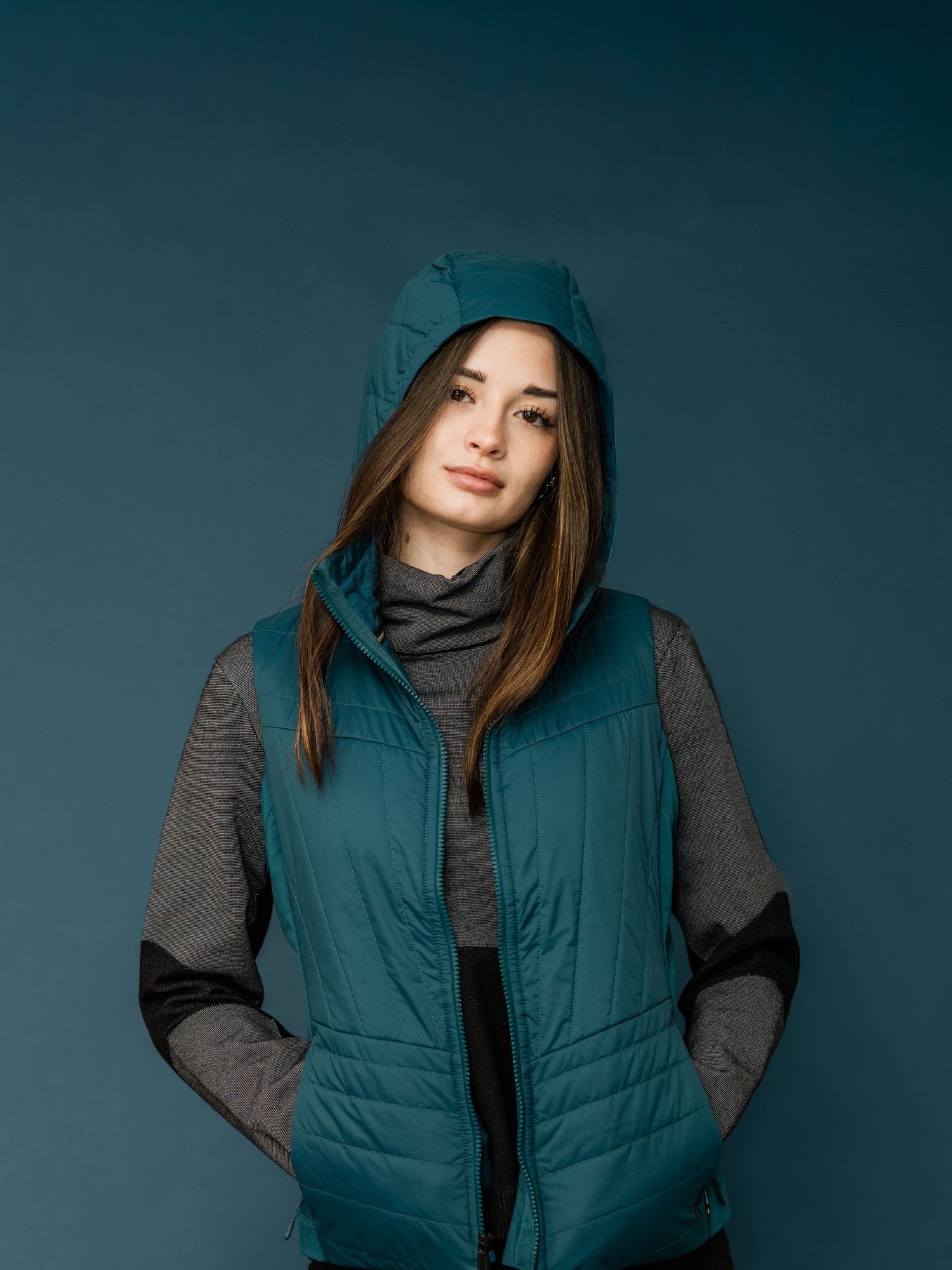 A person wearing a teal puffer vest and gray sweater poses against a dark background in a fashion-style portrait series.