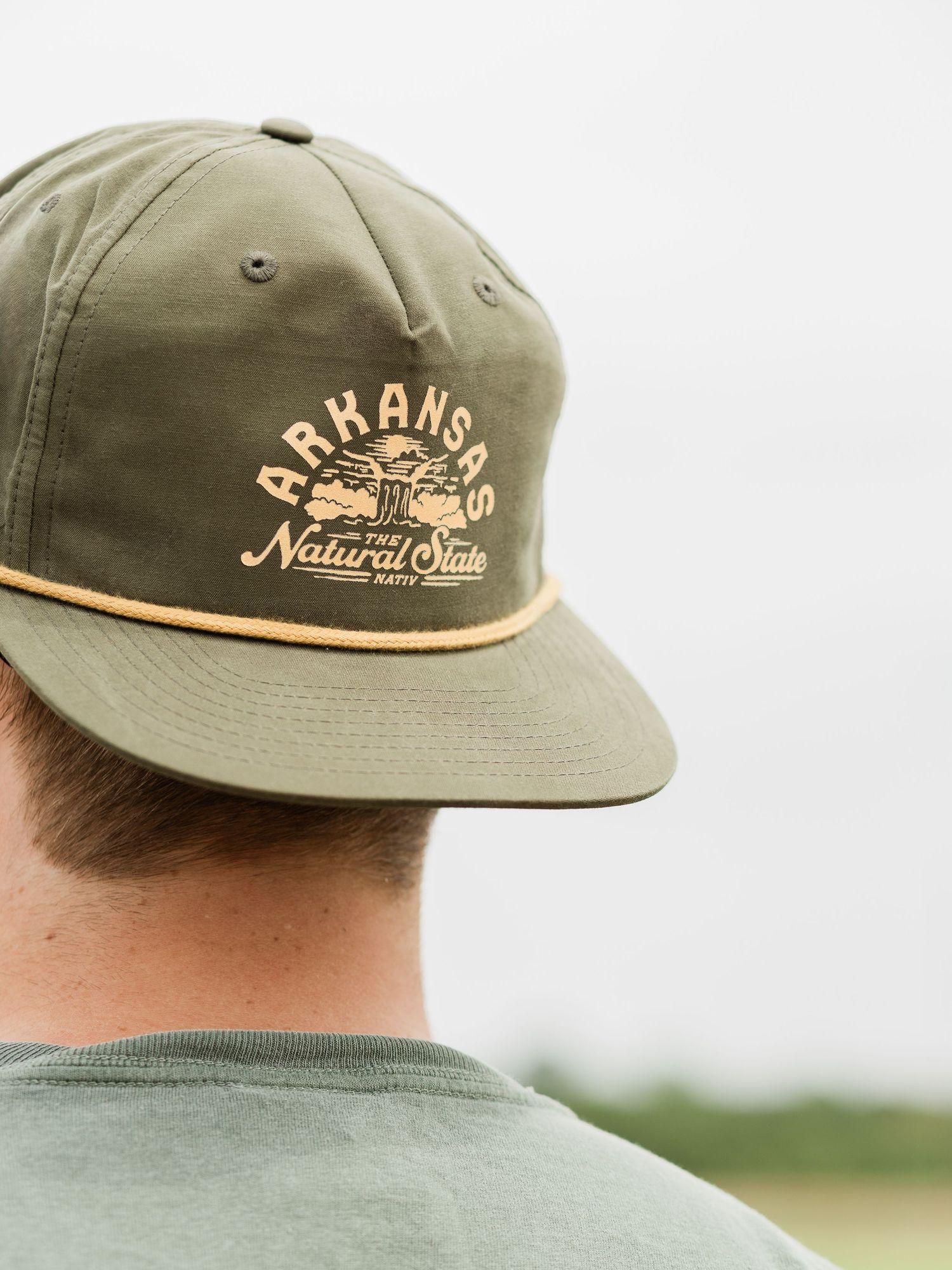 Man wearing an olive green Arkansas The Natural State snapback hat with gold embroidery, viewed from behind.