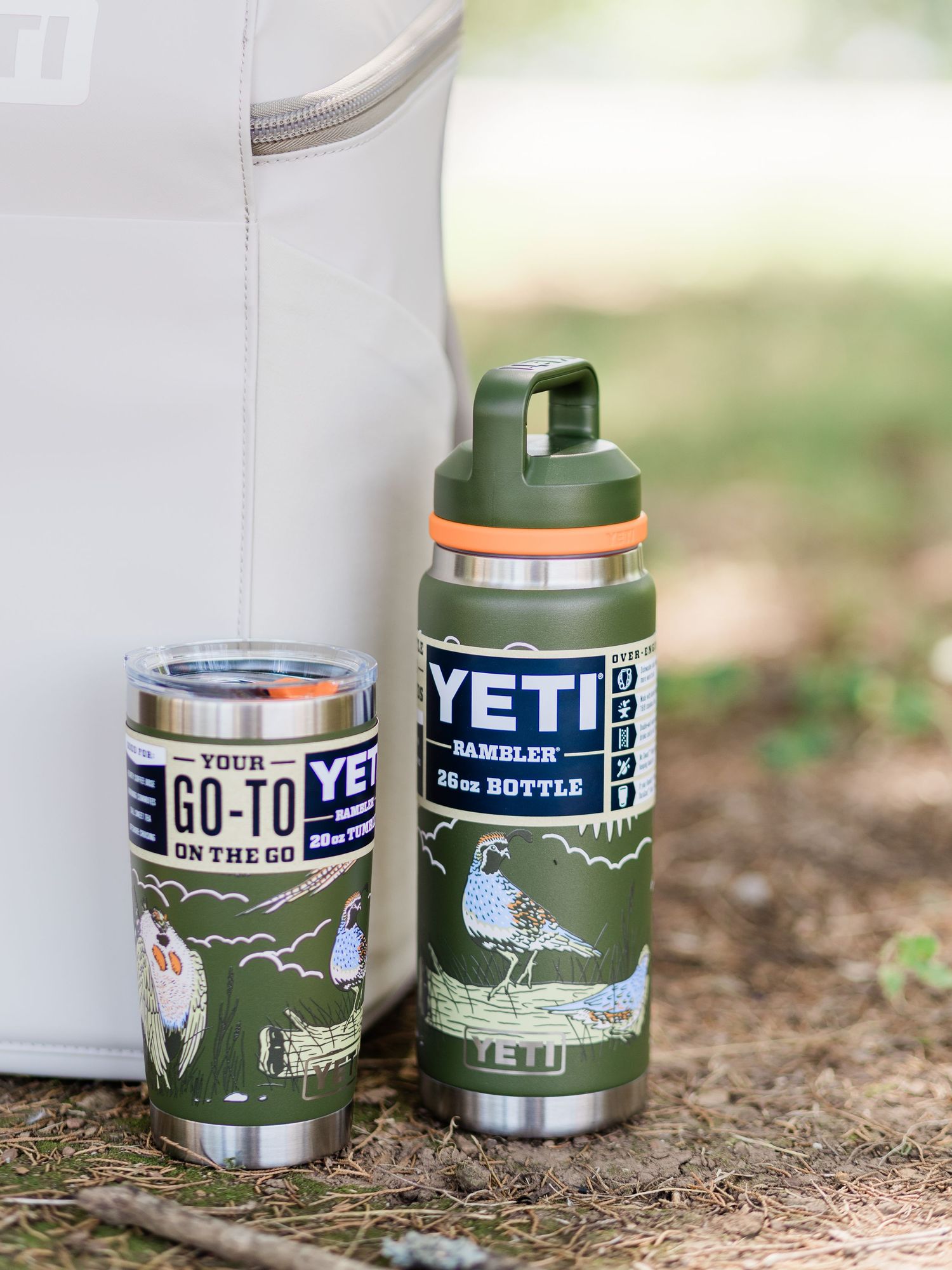 Two olive green YETI Rambler bottles with wildlife graphics sit outdoors against a white cooler bag on natural ground.