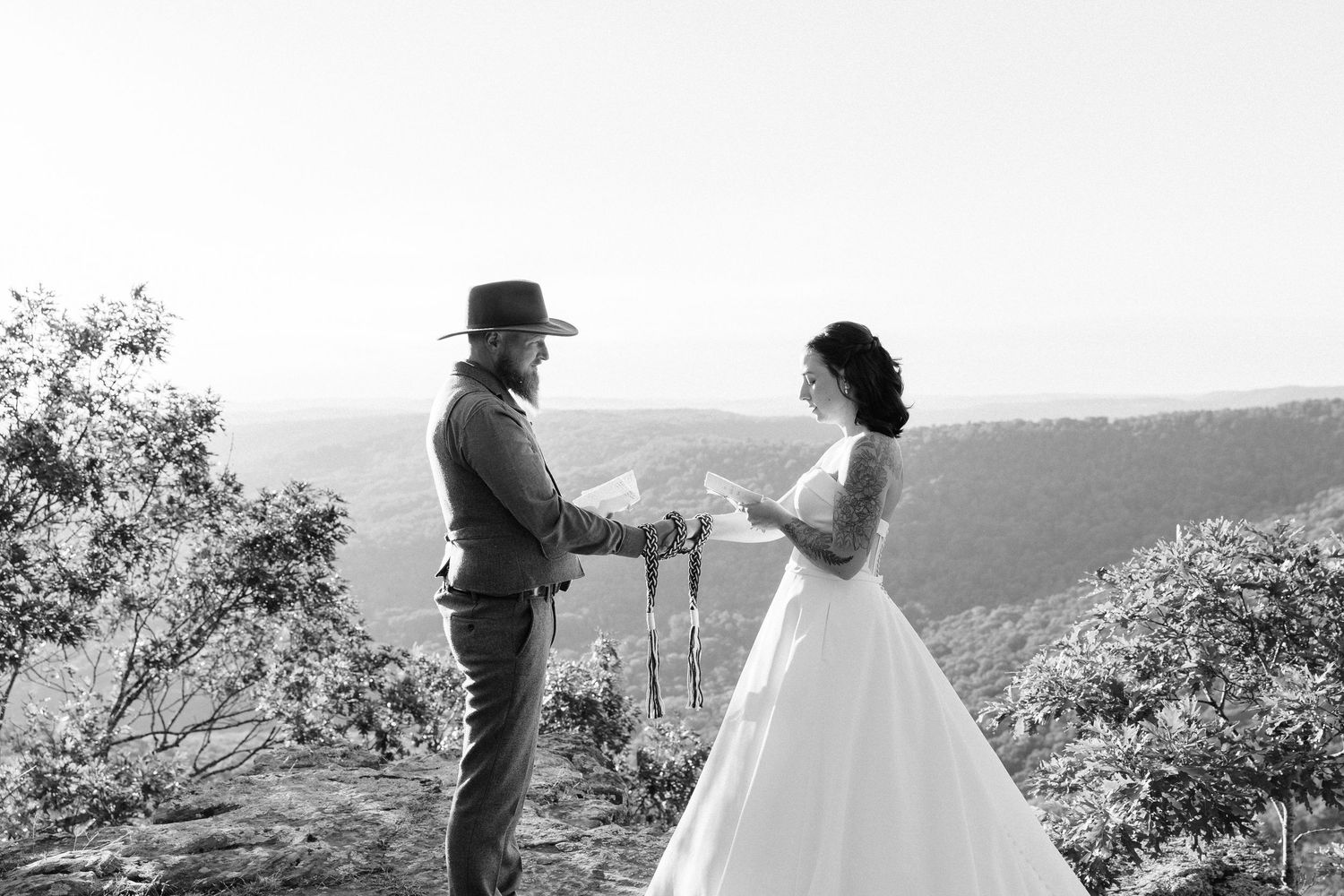 A couple stands in silhouette on a hilltop against a scenic mountain landscape during their wedding photo session.