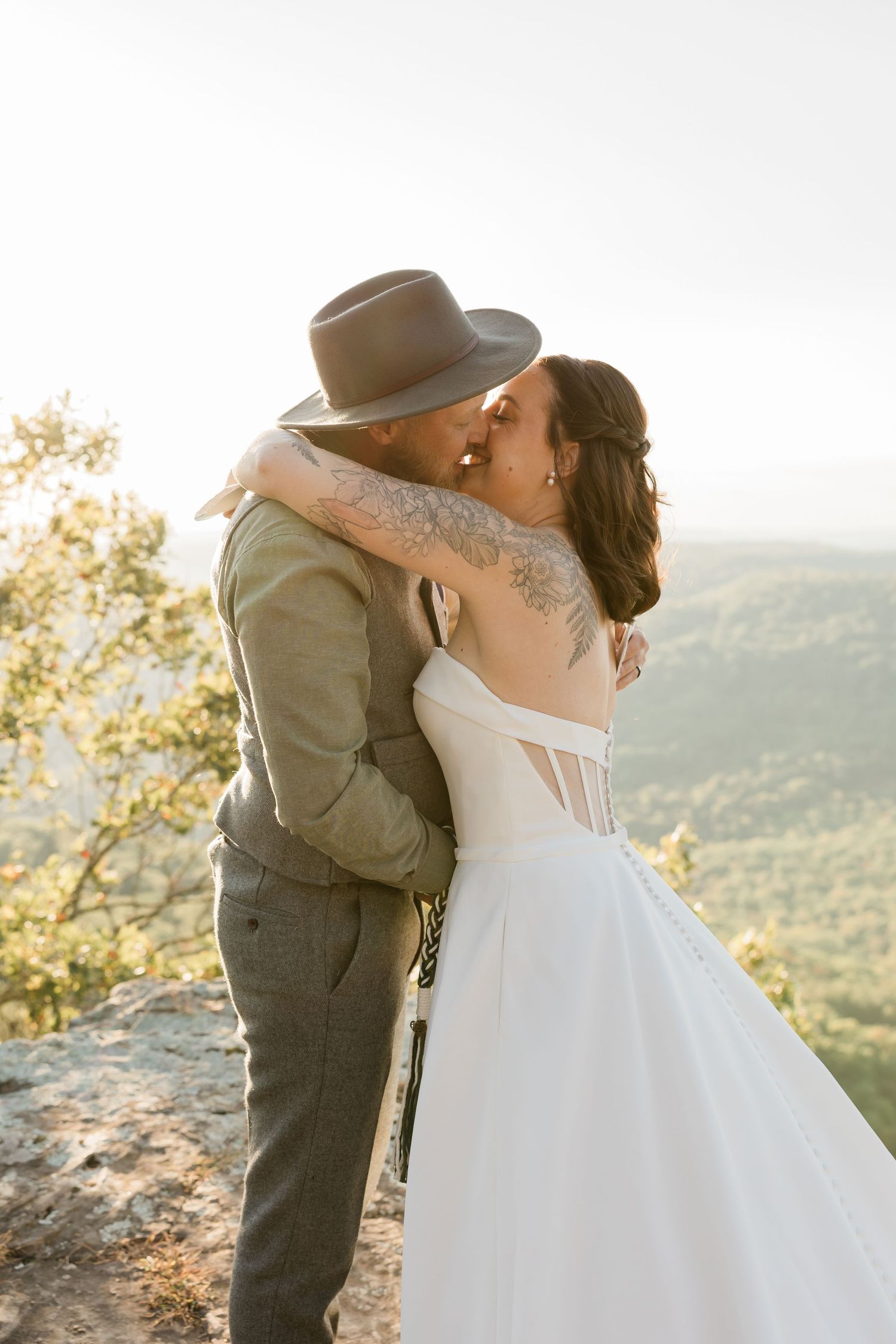 Couple embraces and kisses during sunset on a mountain overlook, bride in white dress and groom in hat and suit.