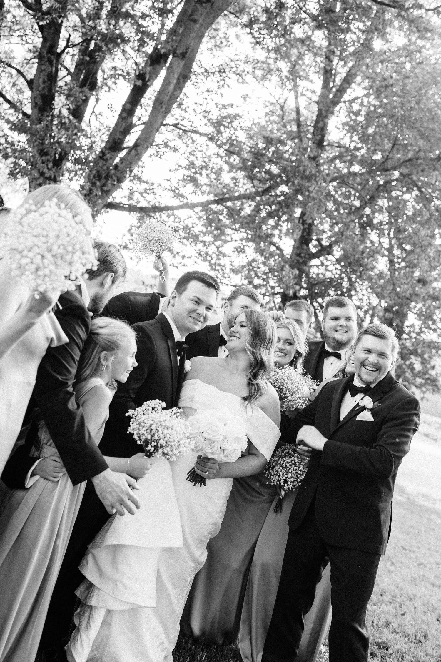 A wedding party laughs together outdoors under trees in a candid black and white photograph.