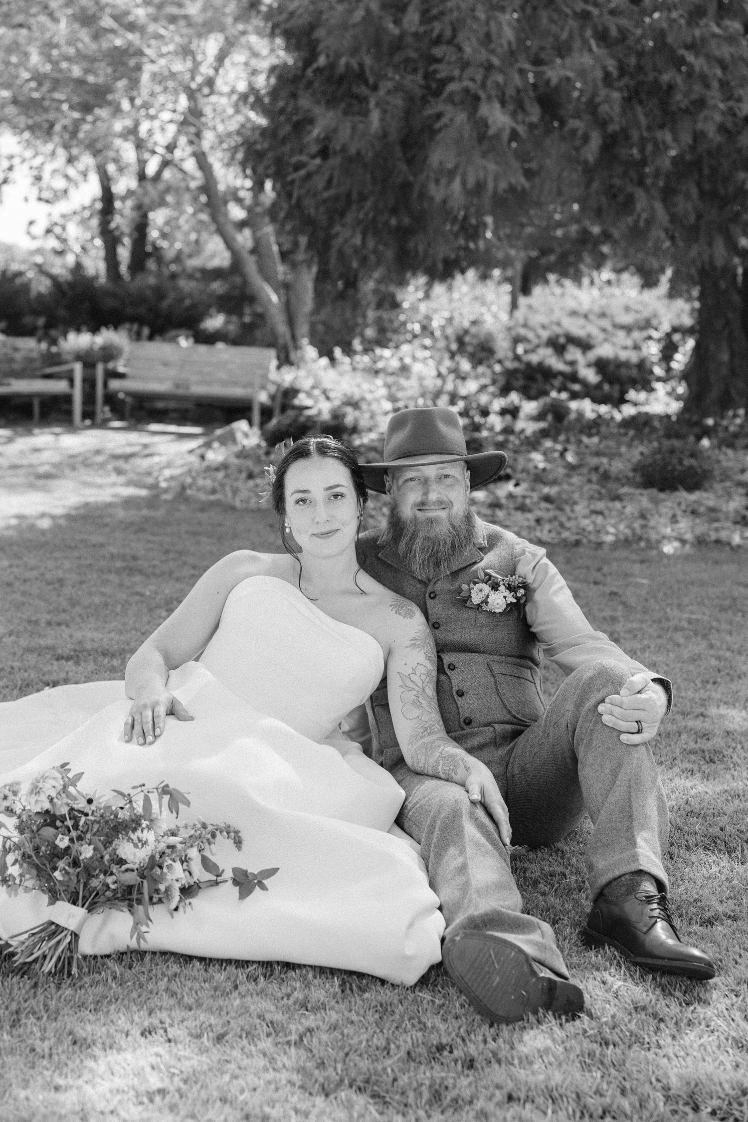 Newlyweds relax on grass during outdoor wedding photo session in a garden setting.