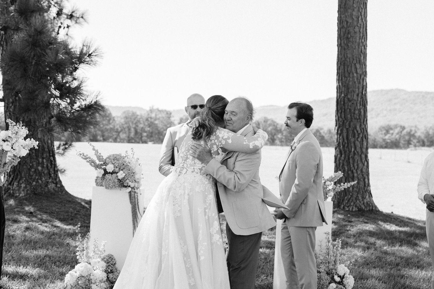 A romantic wedding ceremony takes place outdoors under tall pine trees beside a serene lake.