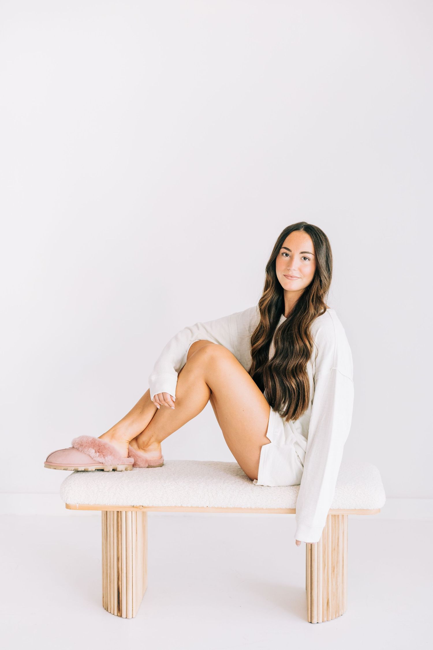 A person in a white sweater sits on a minimalist wooden bench against a white background.