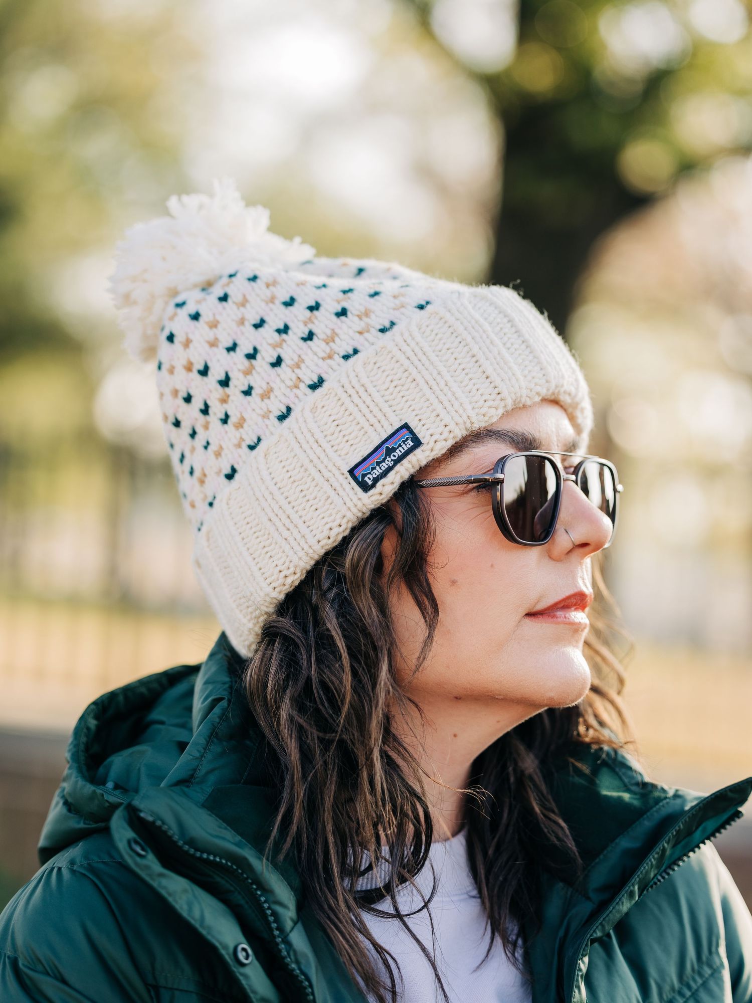A person wearing a white knit beanie with blue polka dots and sunglasses poses outdoors in a dark winter coat.