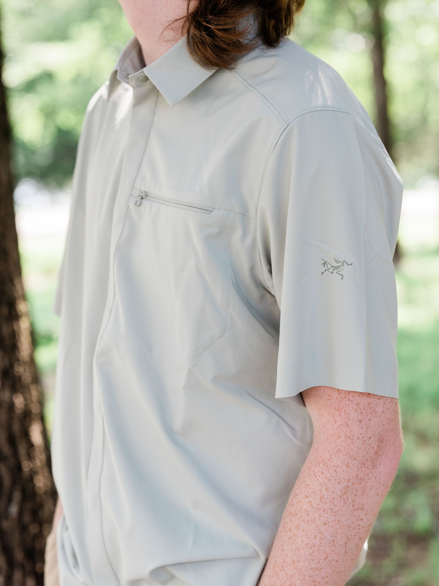 Person wearing a light gray Arc'teryx short-sleeve button-up shirt with chest zip pocket, outdoors.
