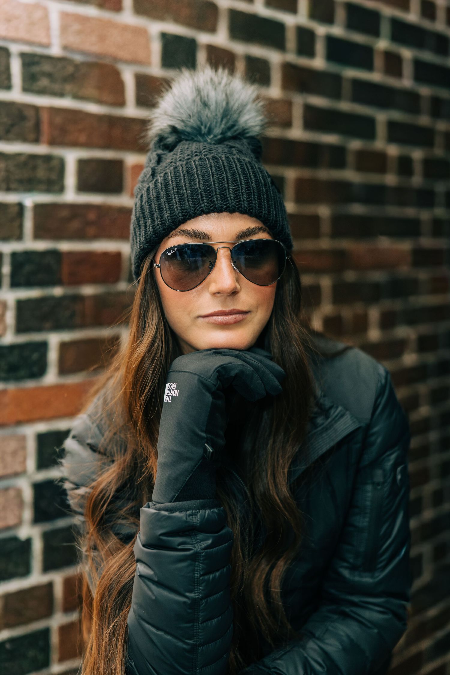 Young woman in gray pom beanie, aviator sunglasses, black gloves and puffer jacket posing against a brick wall.