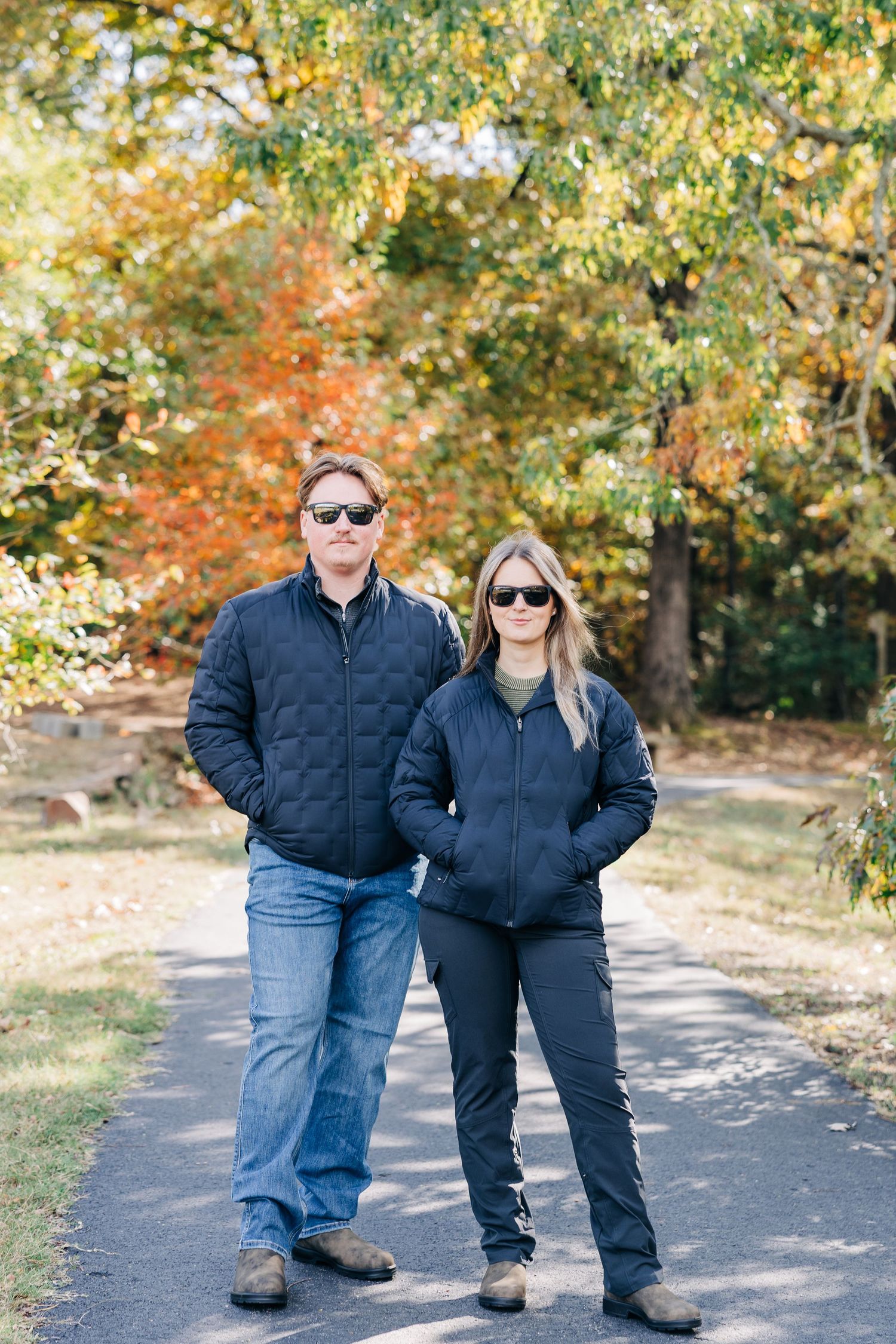 Couple in black jackets and sunglasses standing on a path surrounded by colorful autumn trees.