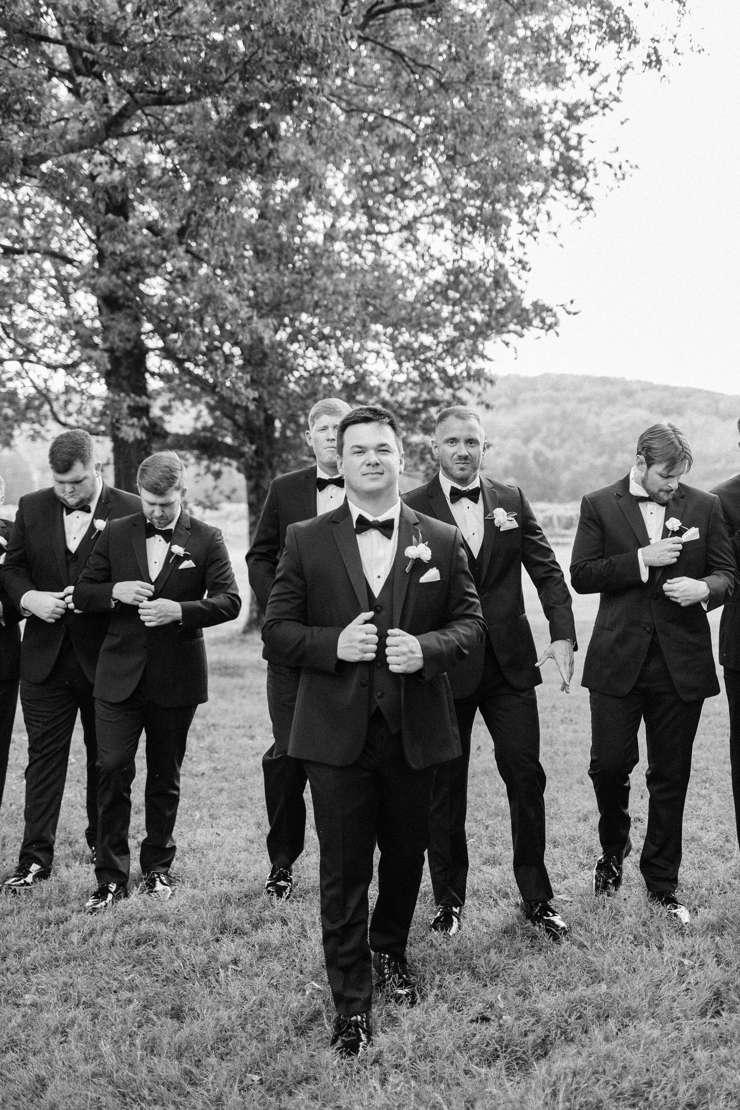 A group of groomsmen in black tuxedos walking together on a grassy hill under a tree in black and white photography.
