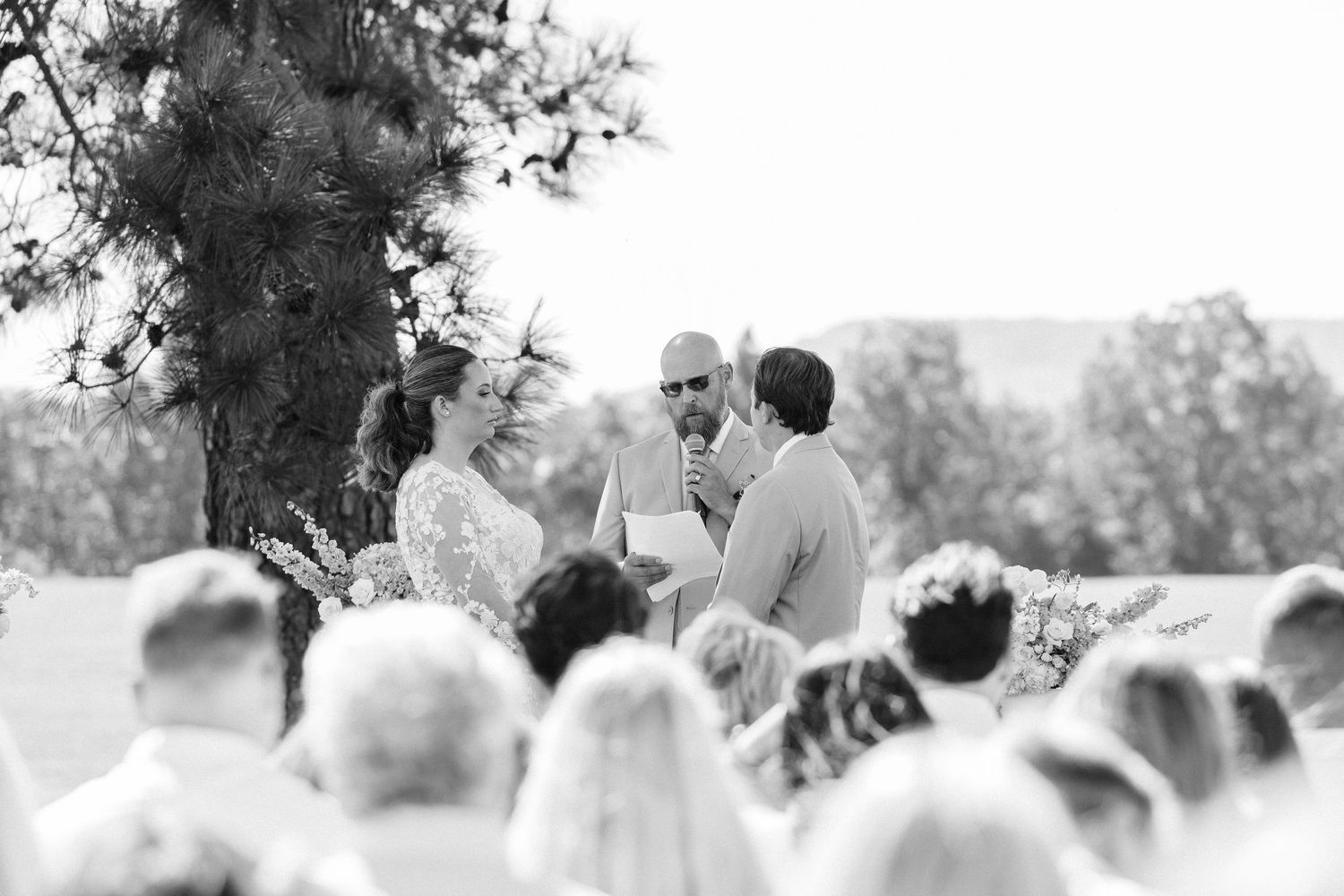 Wedding ceremony taking place on a grassy hillside under tall trees.