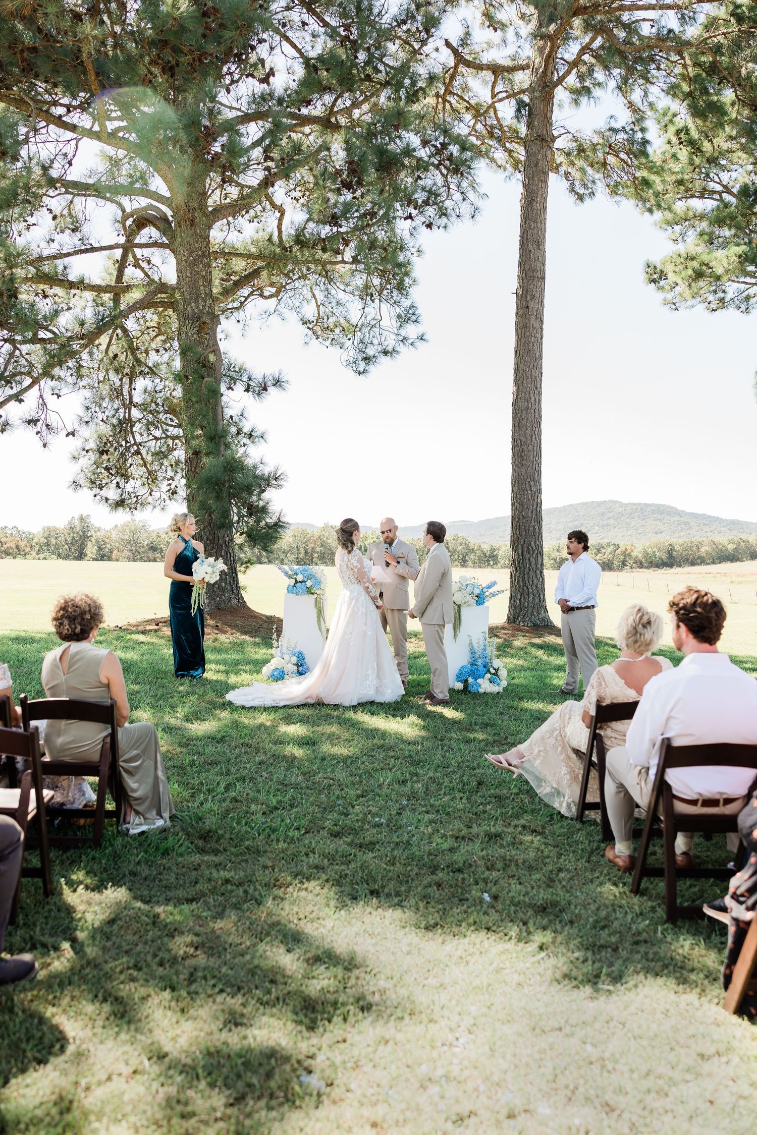 Outdoor wedding ceremony setup under tall pine trees with white chairs and natural lighting.