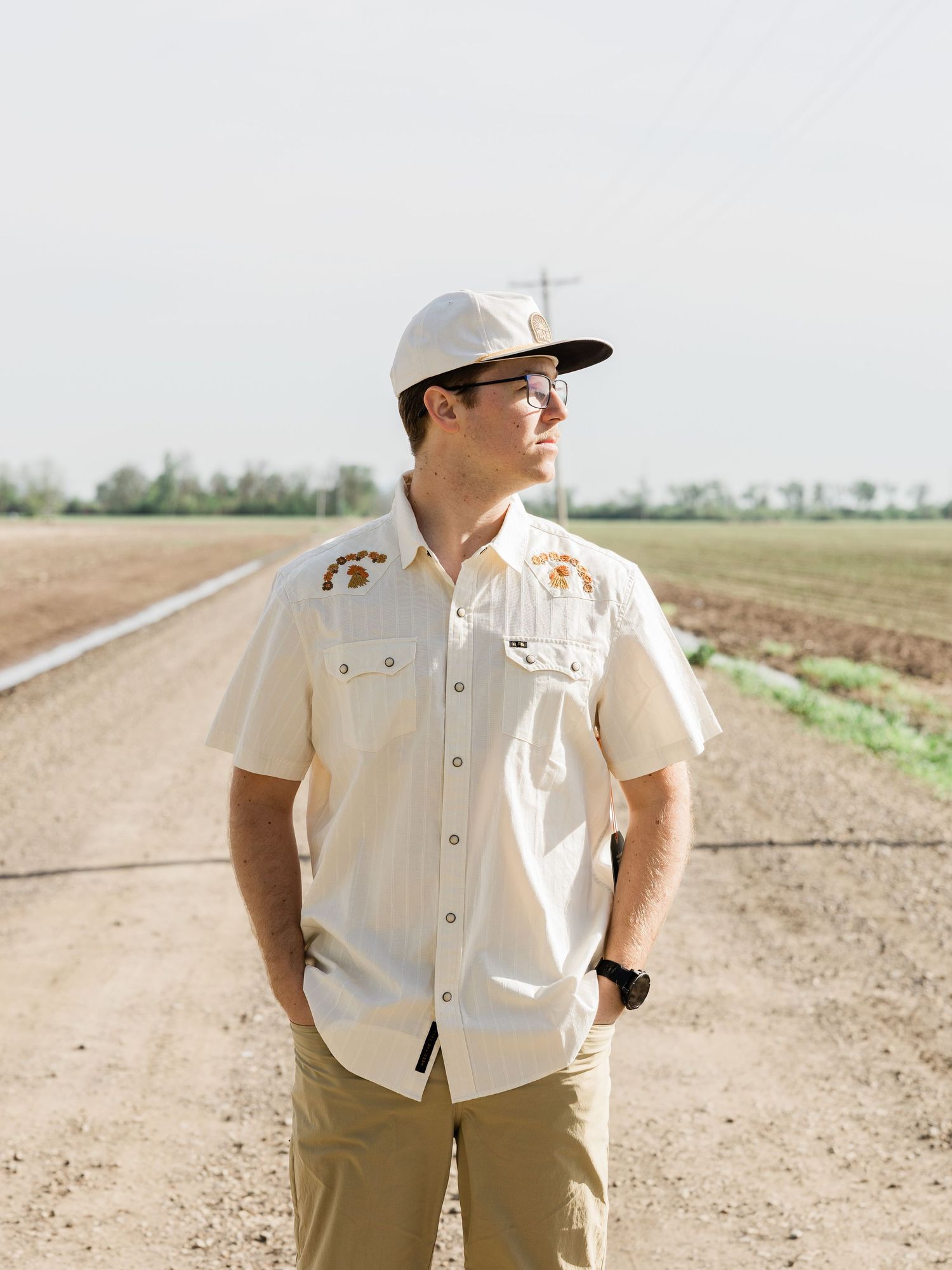 Young man in cream shirt and cap stands on dirt farm road surrounded by agricultural fields.