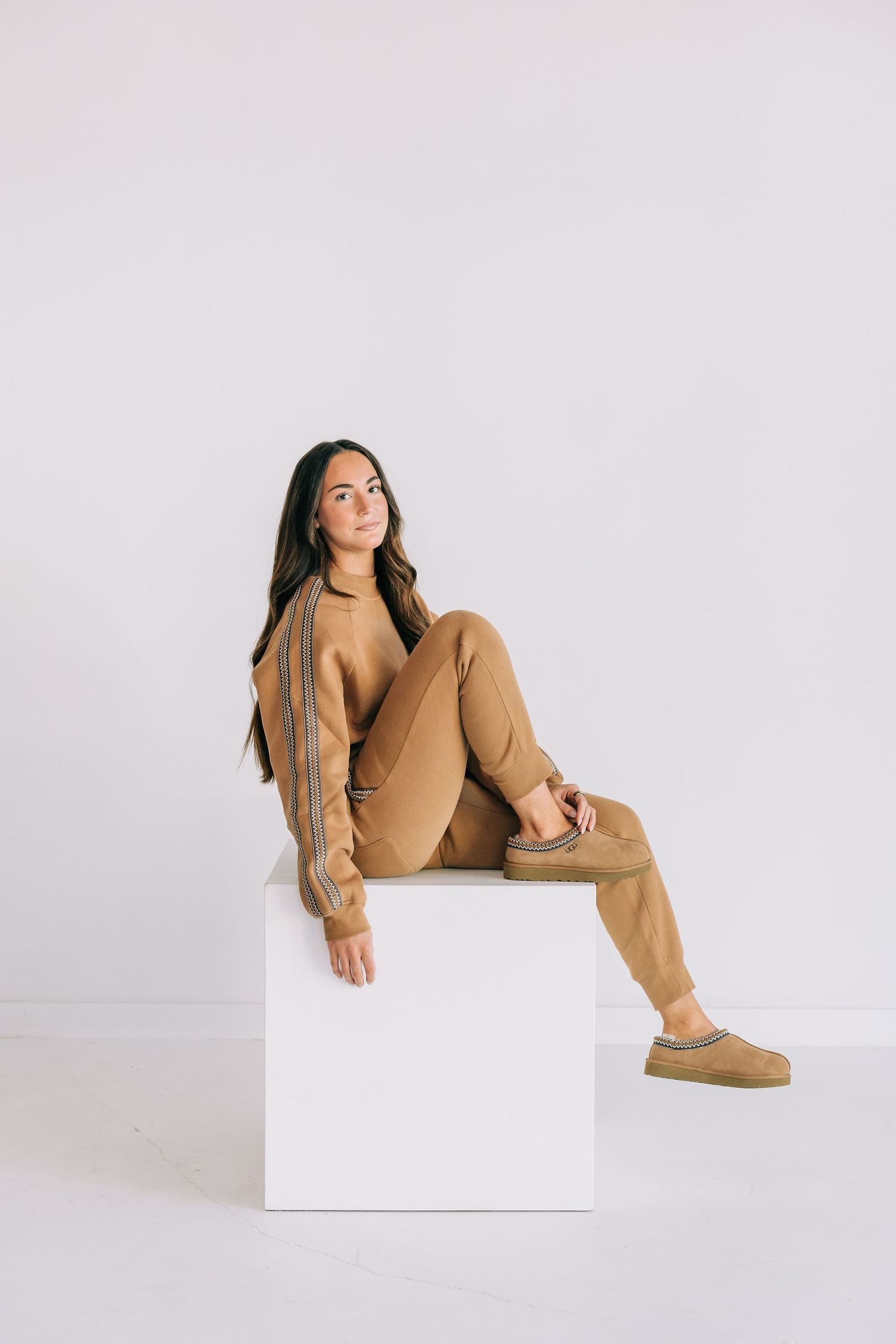 Woman in tan matching tracksuit and UGG slippers seated on white cube against light background.