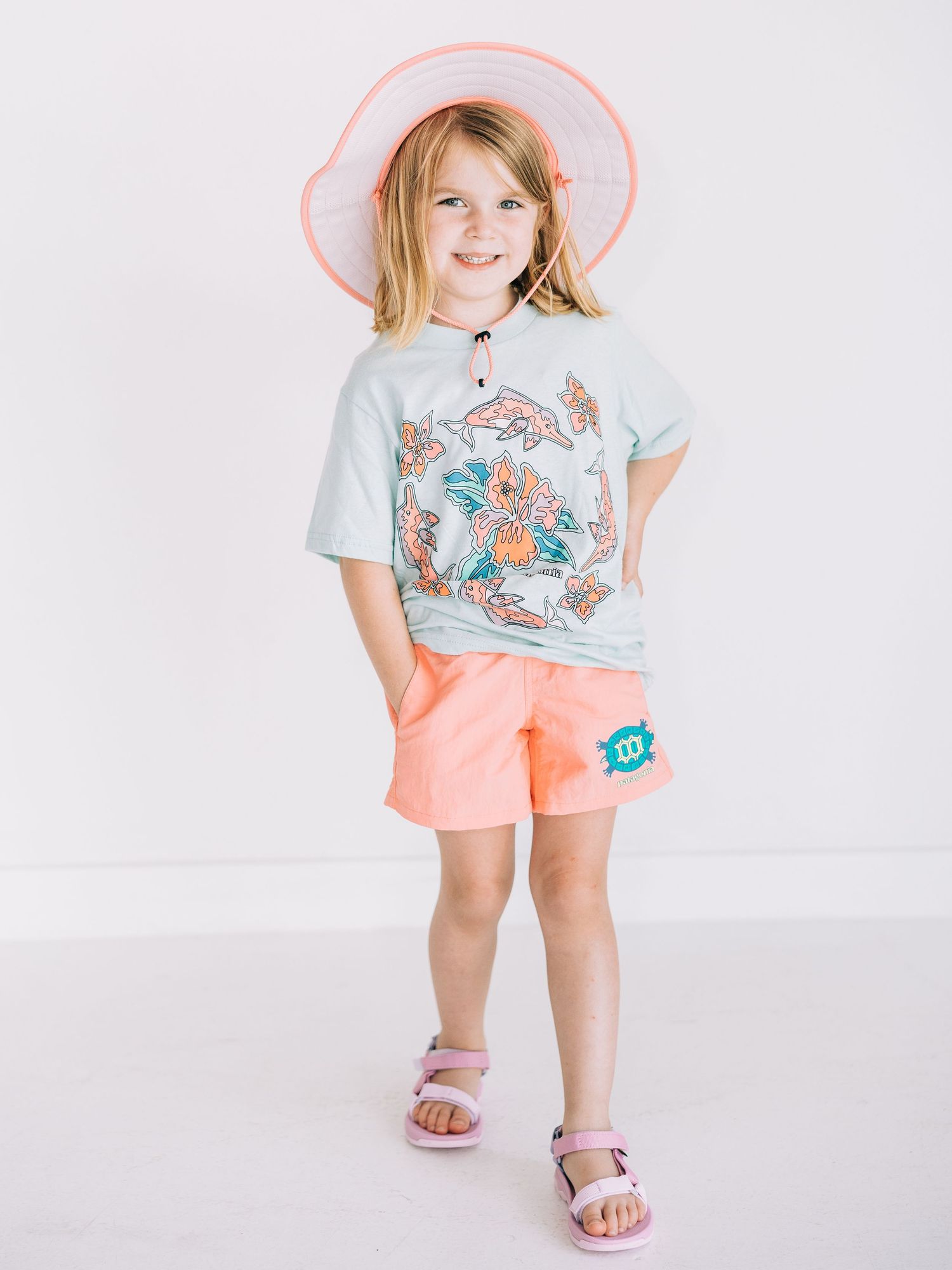 Young girl wearing a mint graphic tee, coral shorts, pink sun hat, and lavender sandals poses against white background.