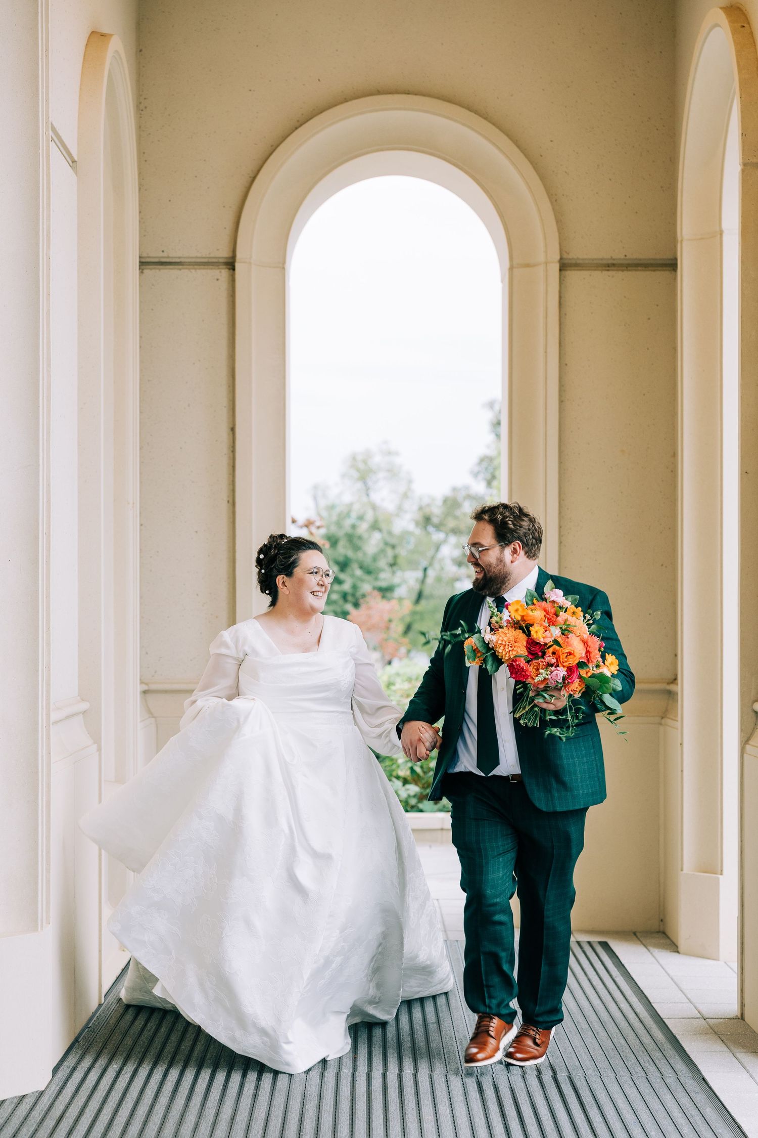 Couple walks through arched hallway, bride in white dress and groom in emerald suit.