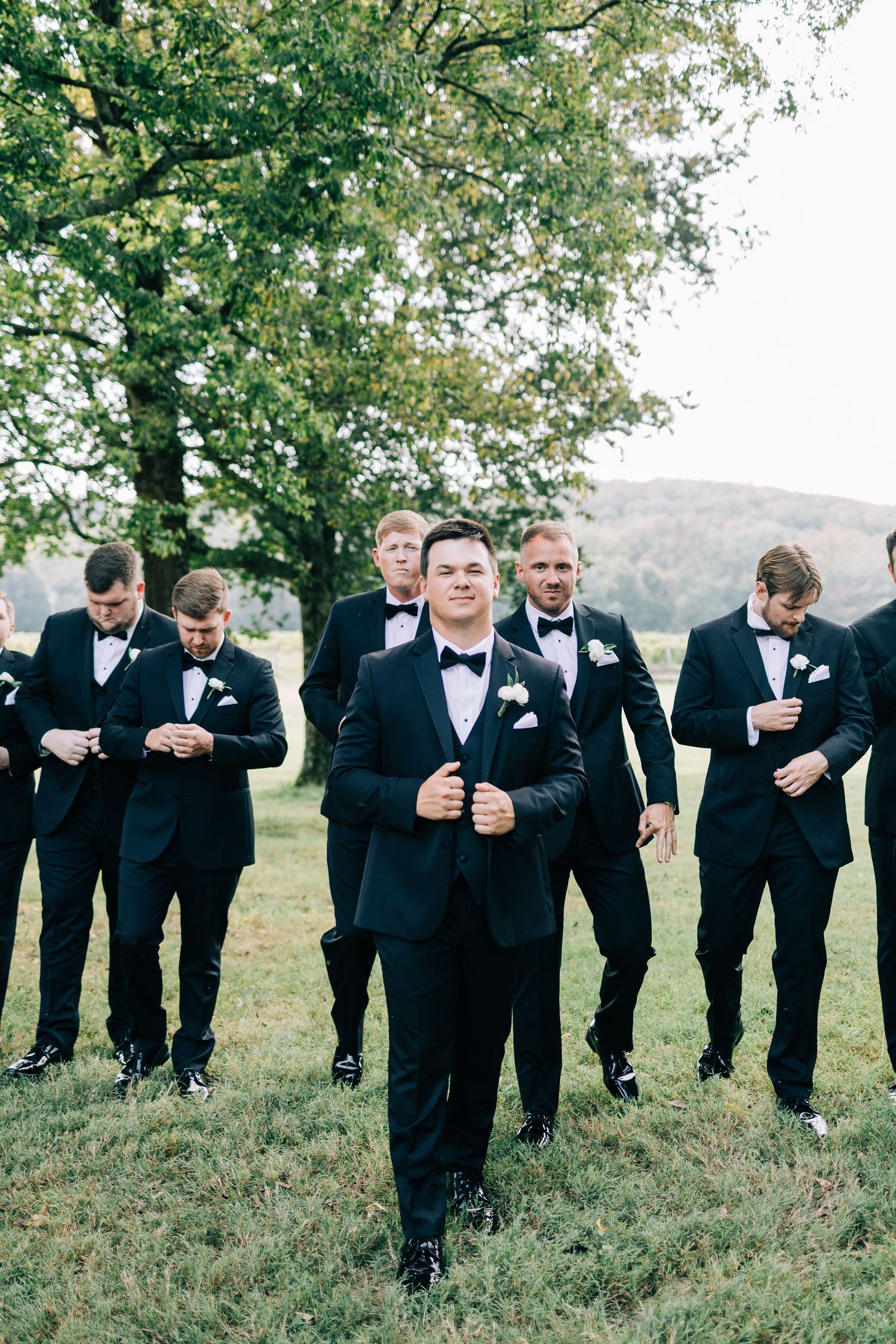 Groomsmen in black tuxedos pose in a casual walking formation on grassy terrain under trees.