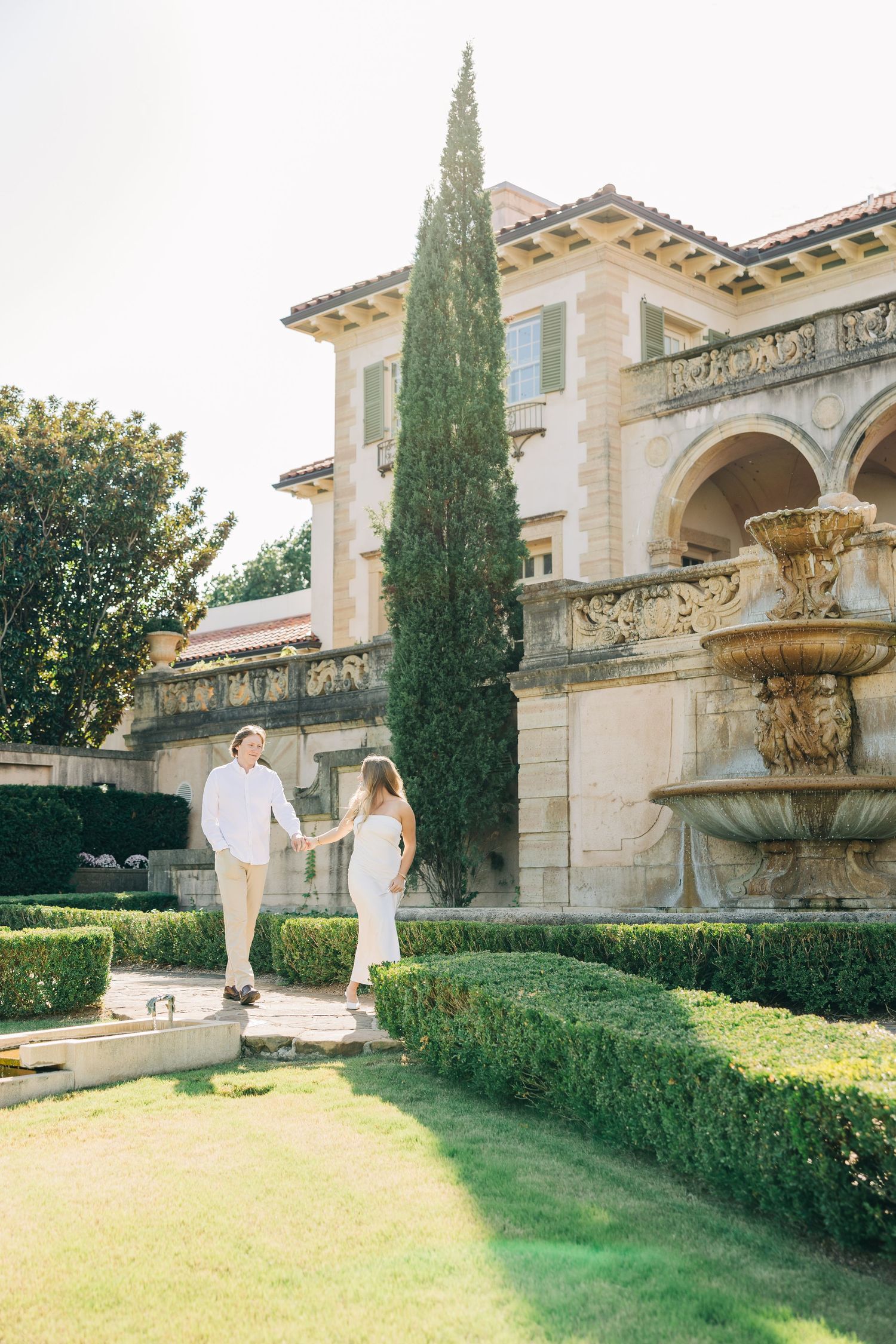 A couple strolls through manicured gardens beside an elegant Mediterranean-style mansion with a decorative fountain.