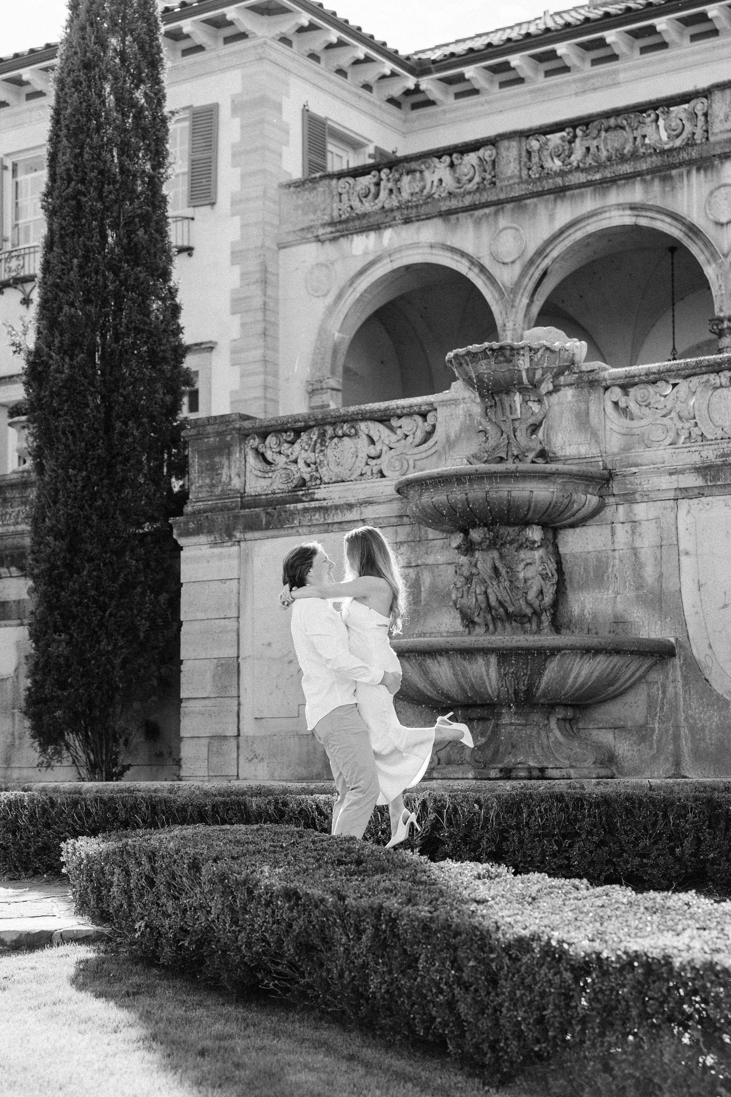 A figure in white performs graceful dance poses in a garden setting with stone architecture and cypress trees.