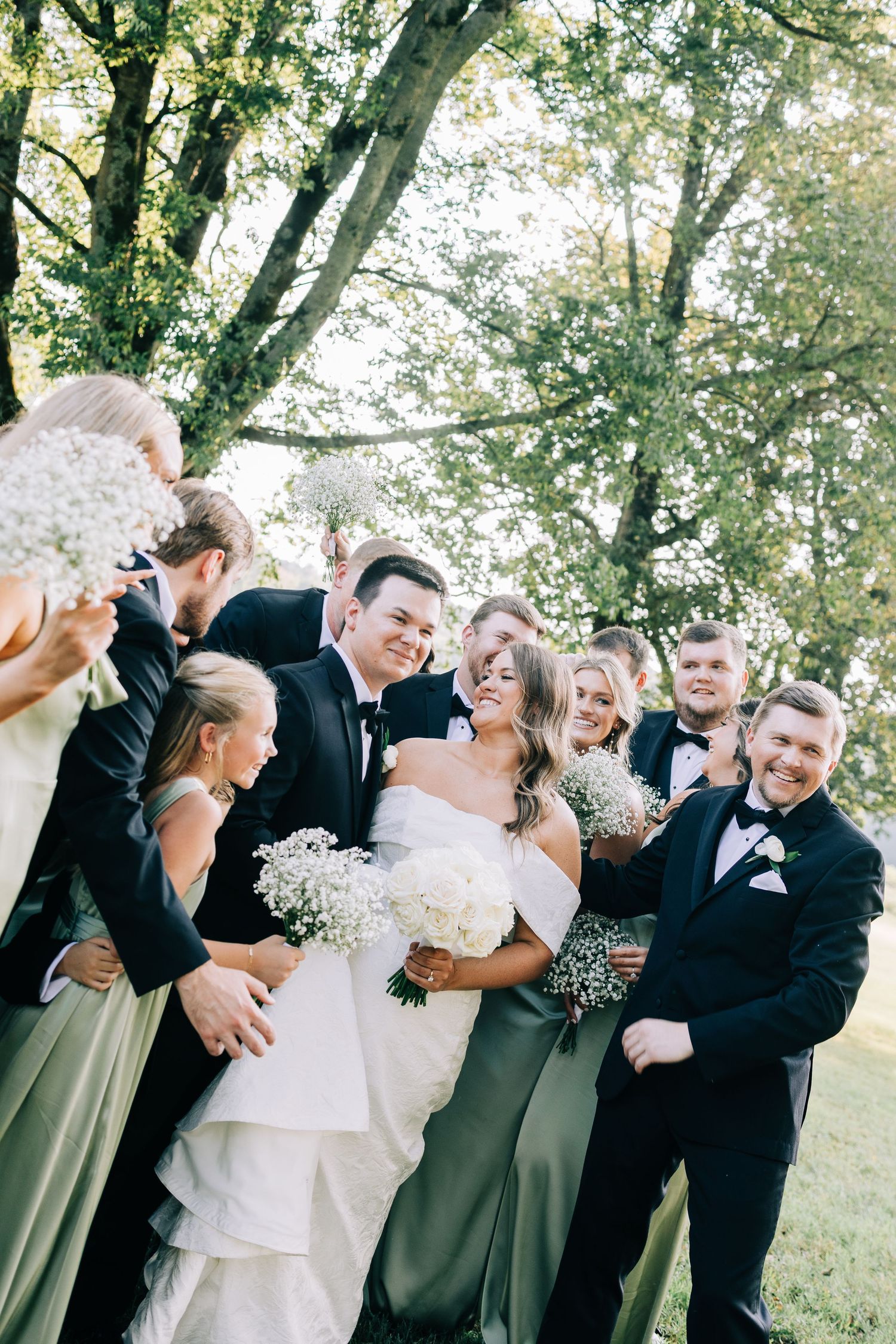 Wedding party in formal attire shares joyful candid moments outdoors under green trees.