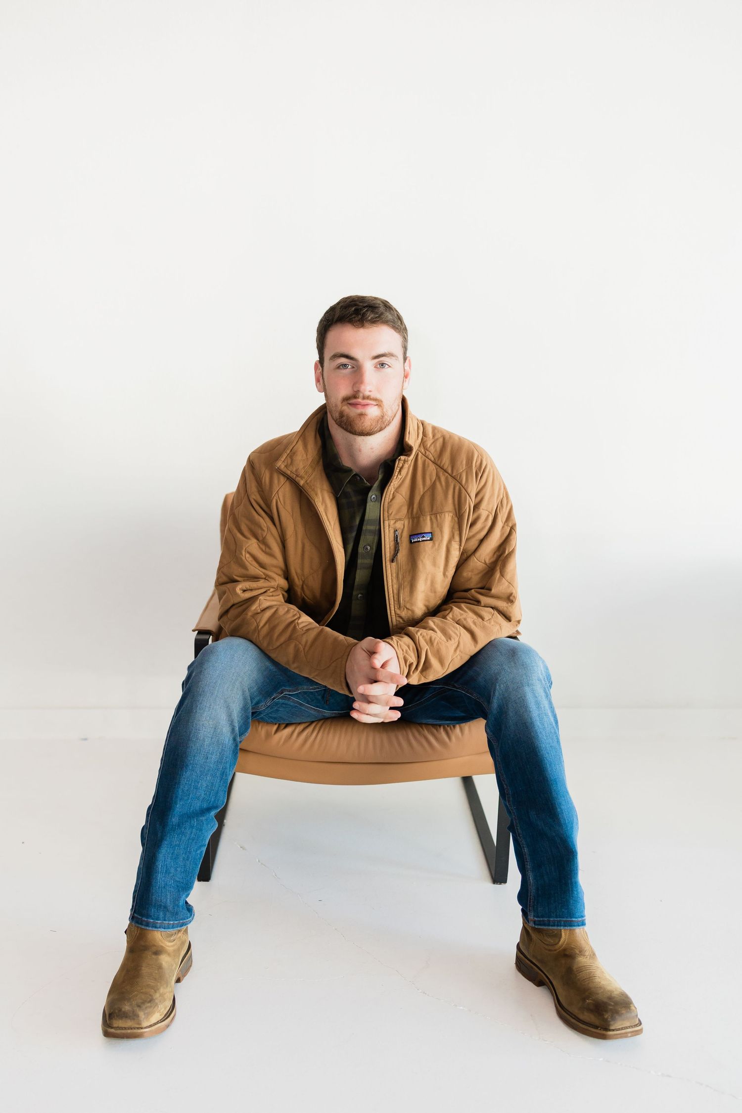 Person wearing brown jacket and jeans sits in wide stance on white background in studio setting.