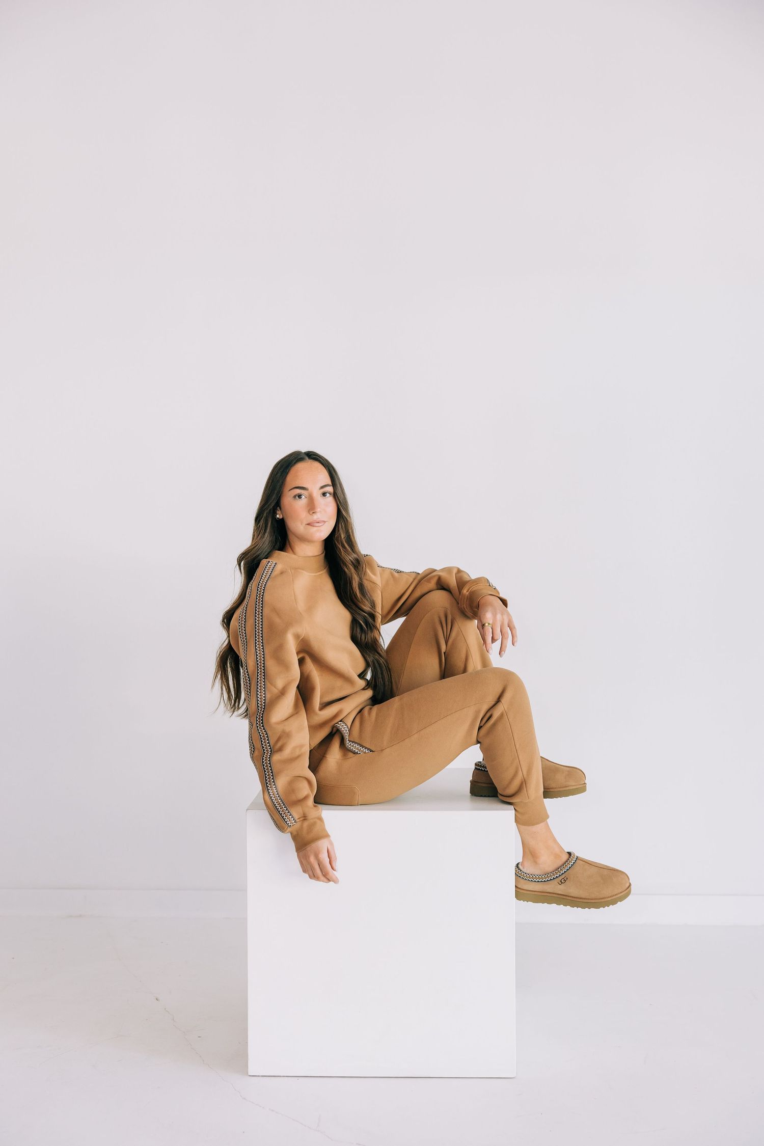 Person in tan loungewear strikes four different casual poses on white background while sitting on white cube.