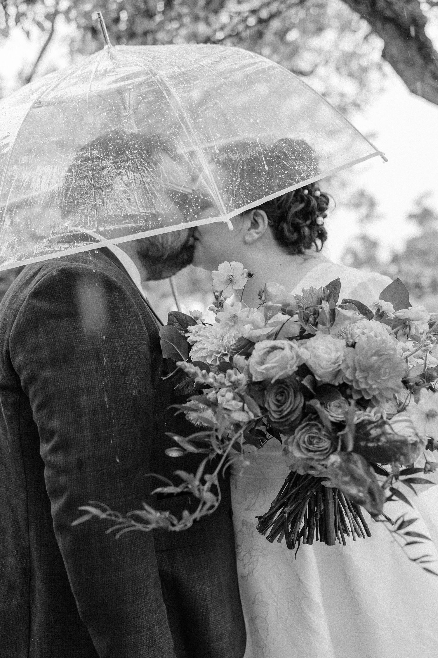 A black and white wedding photo of a couple sharing a moment under a clear umbrella with floral bouquet.
