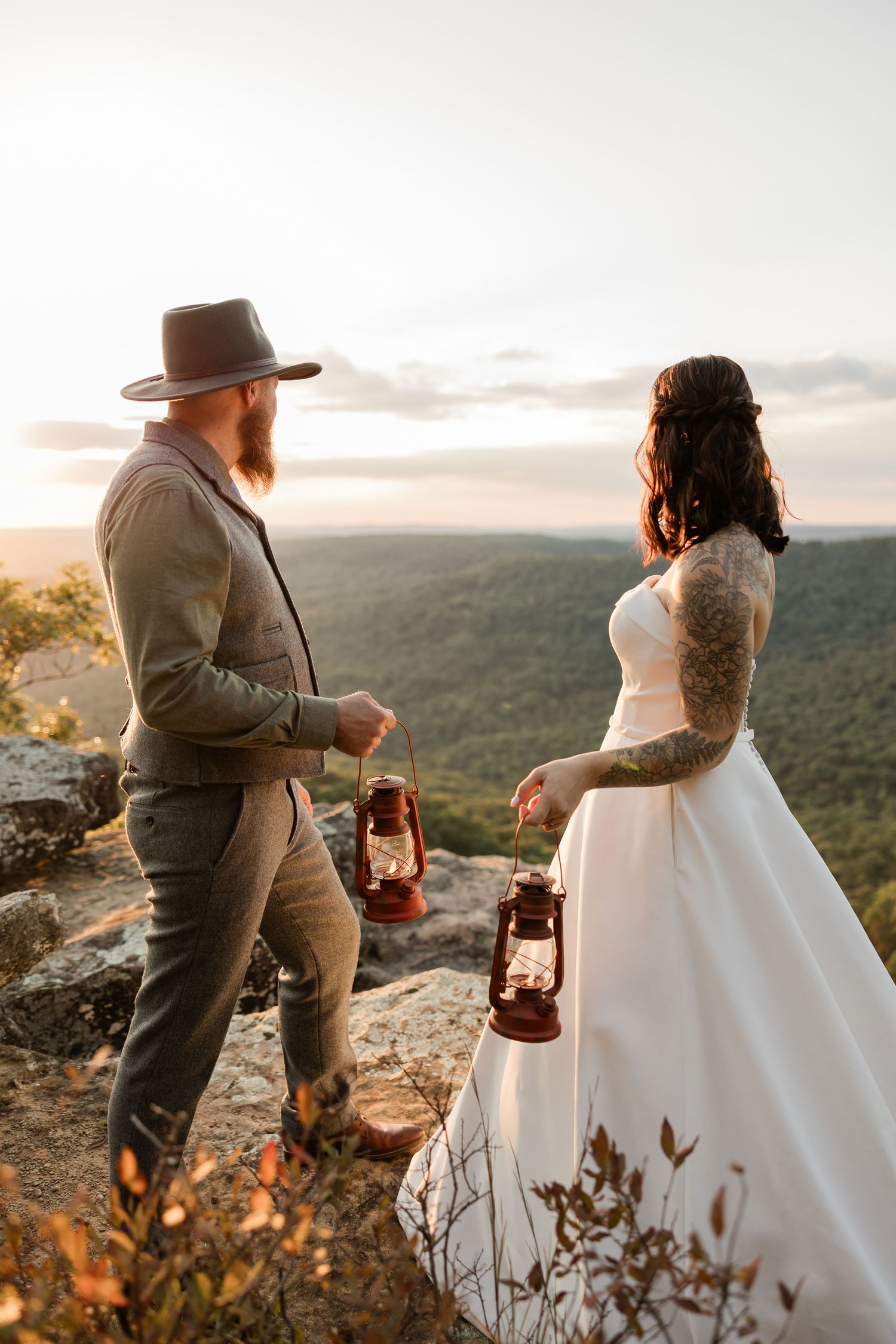 A couple in wedding attire shares an intimate moment on a mountain cliff with a vintage lantern.