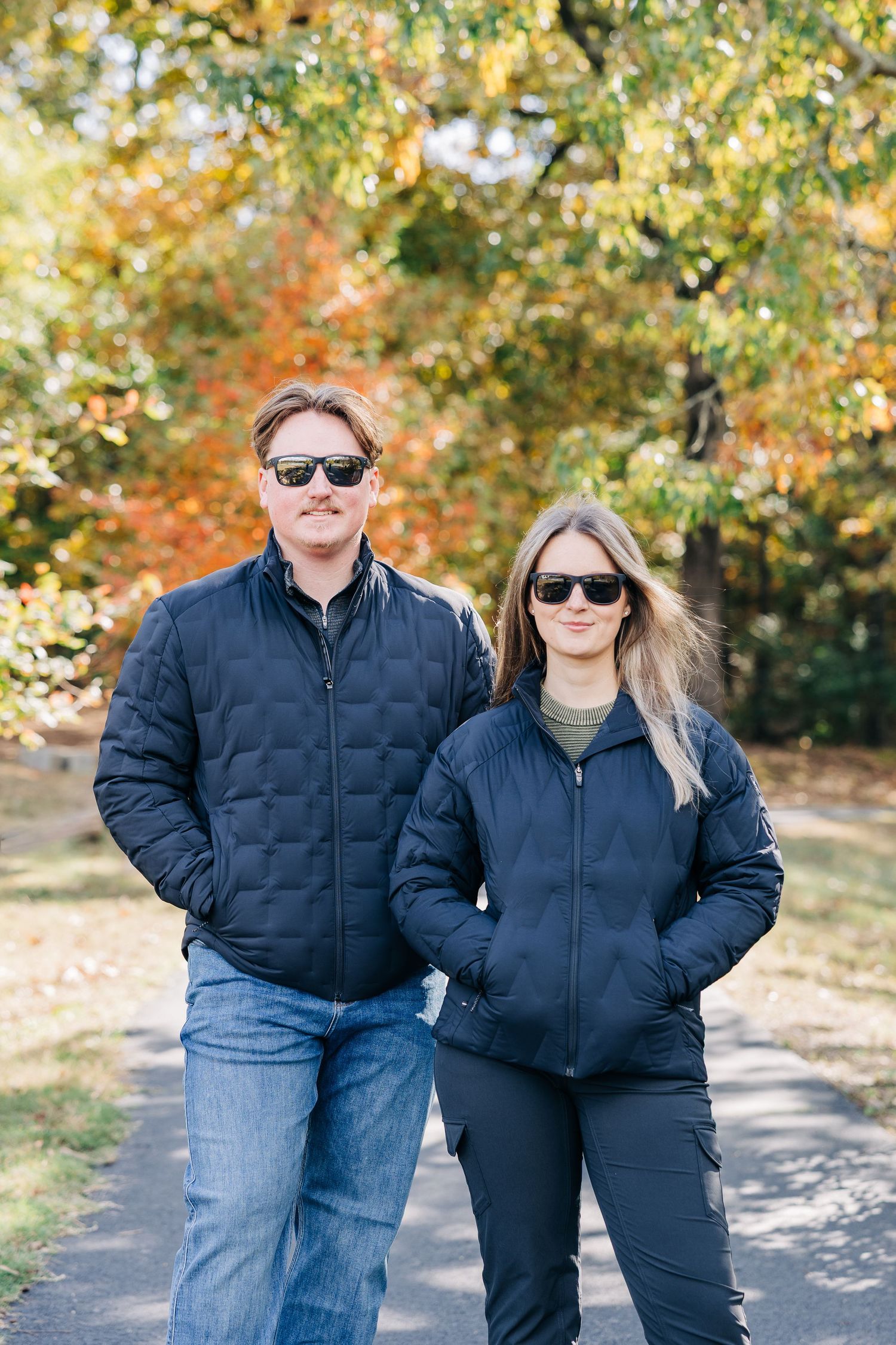 A man and woman in matching navy puffer jackets and sunglasses walk together on a path surrounded by autumn foliage.