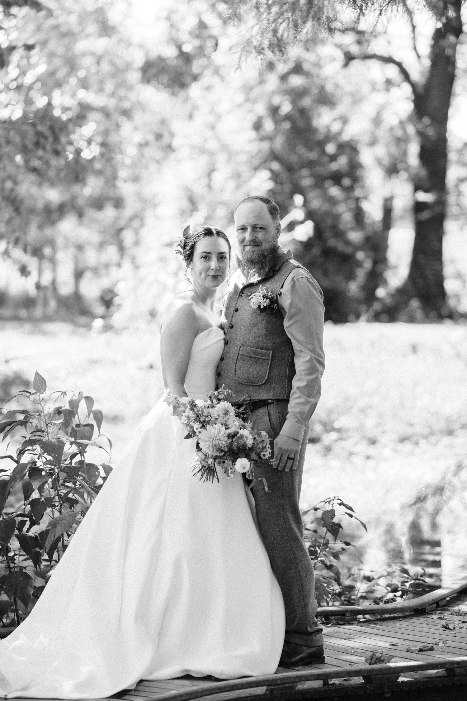 Black and white wedding photography shows couple posing outdoors with floral bouquet in natural setting.