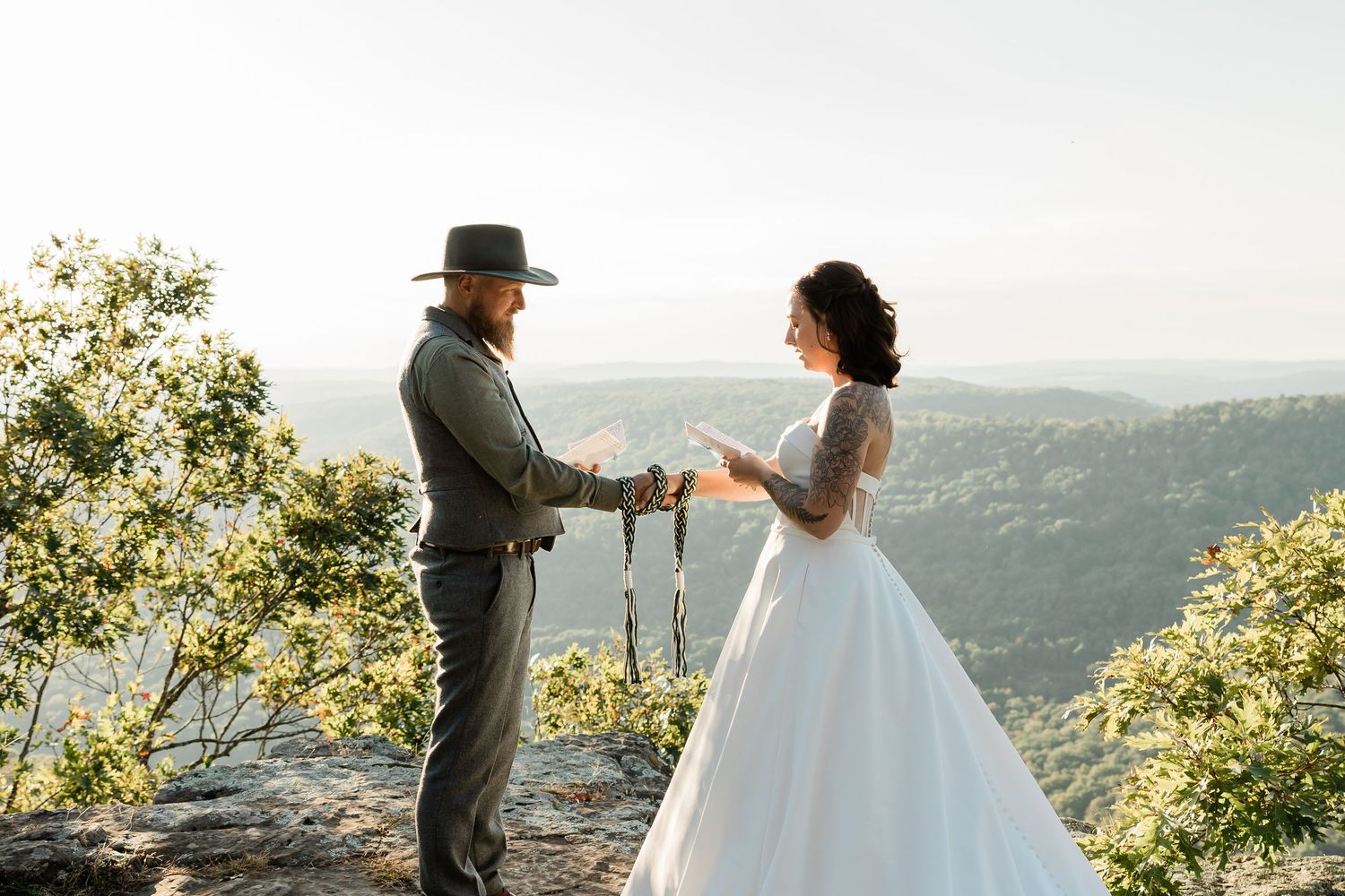 A couple in wedding attire stands together on a scenic mountain overlook during golden hour.