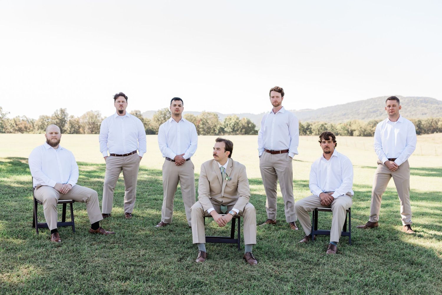 Groomsmen in light colored suits sit casually on a grassy hillside.