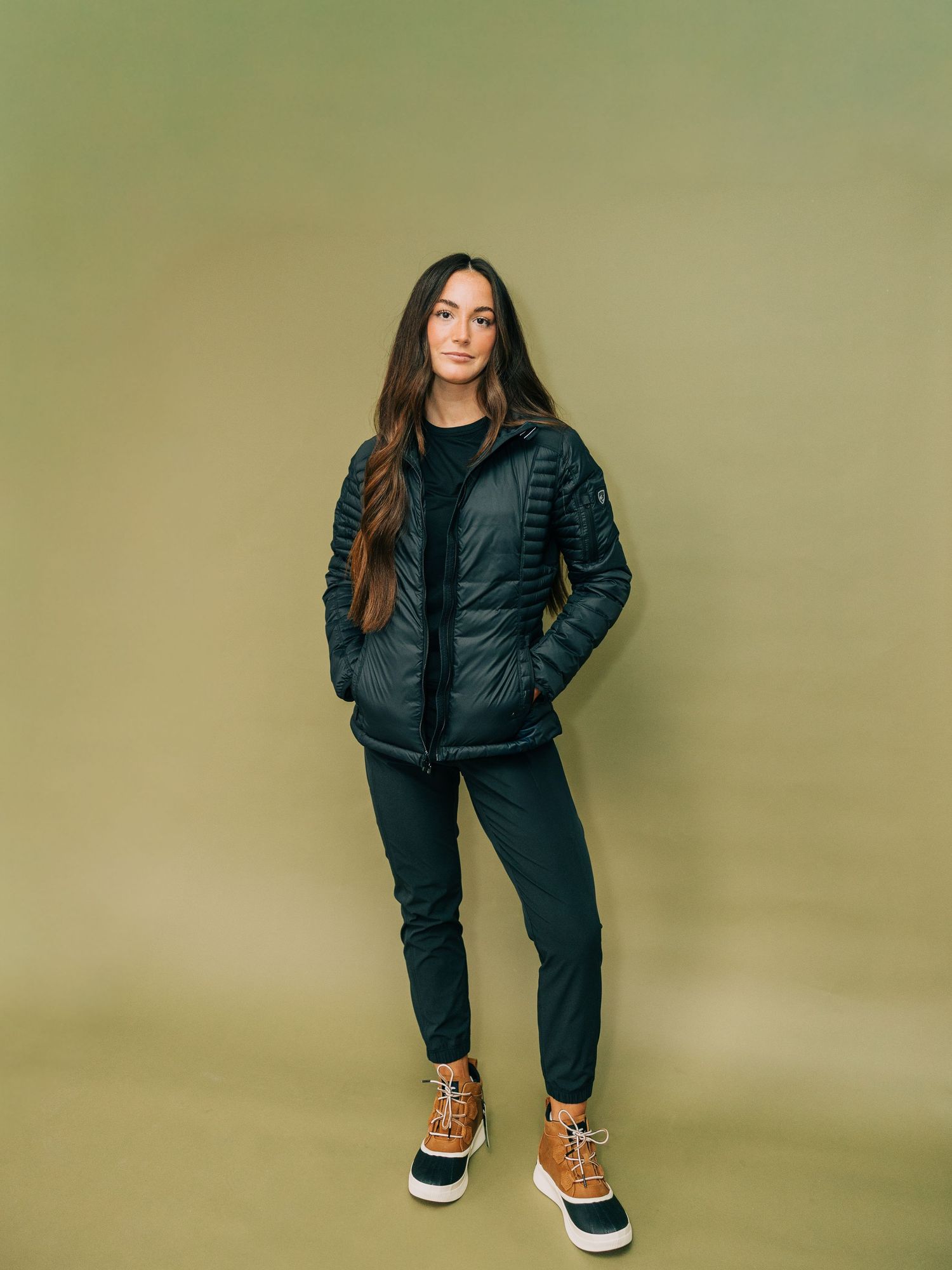 Woman wearing a black puffer jacket, dark joggers, and tan snow boots, posed against an olive green background.
