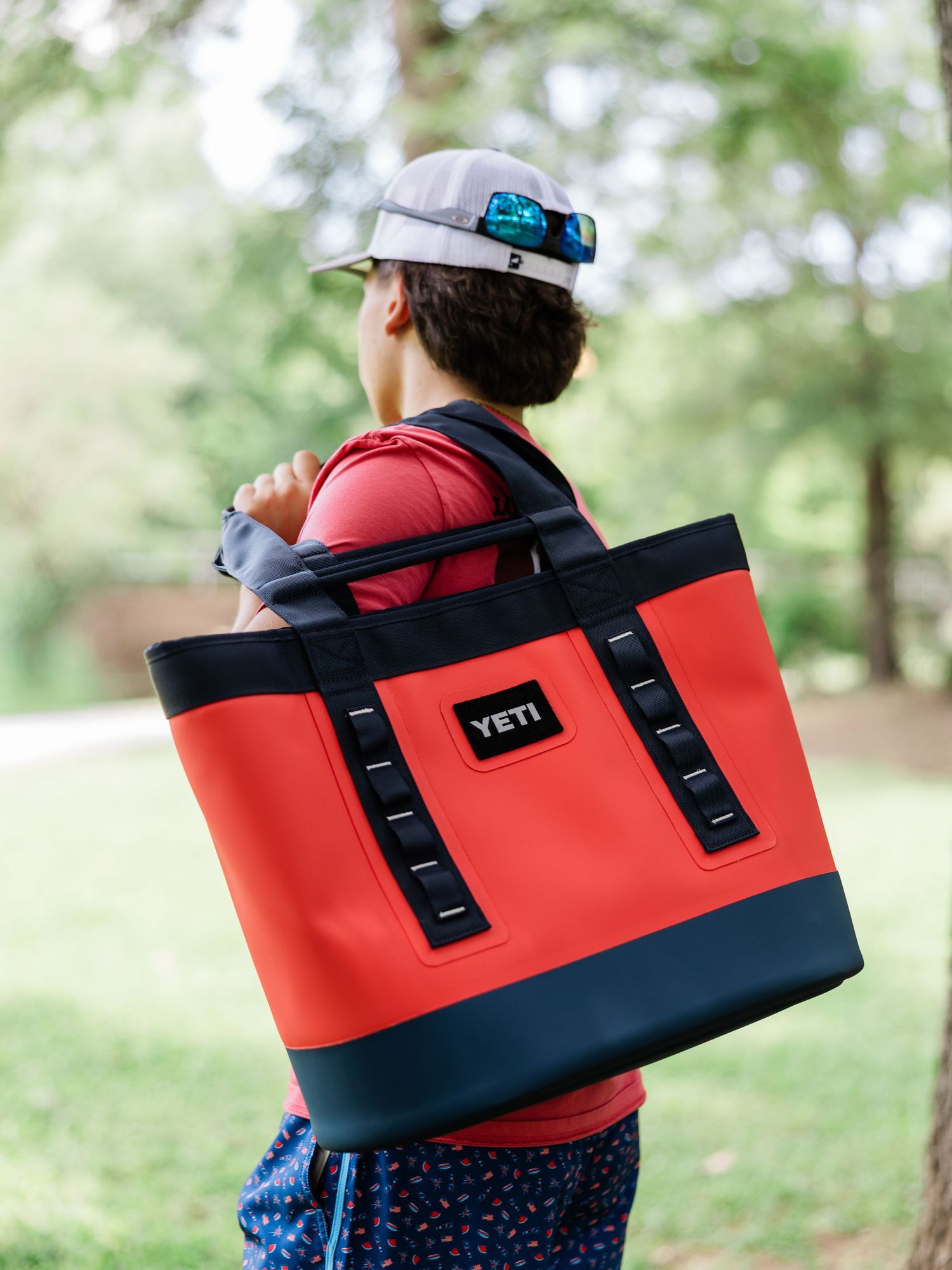 Person carrying a red and navy YETI Camino tote bag outdoors, wearing a cap with sunglasses on top.